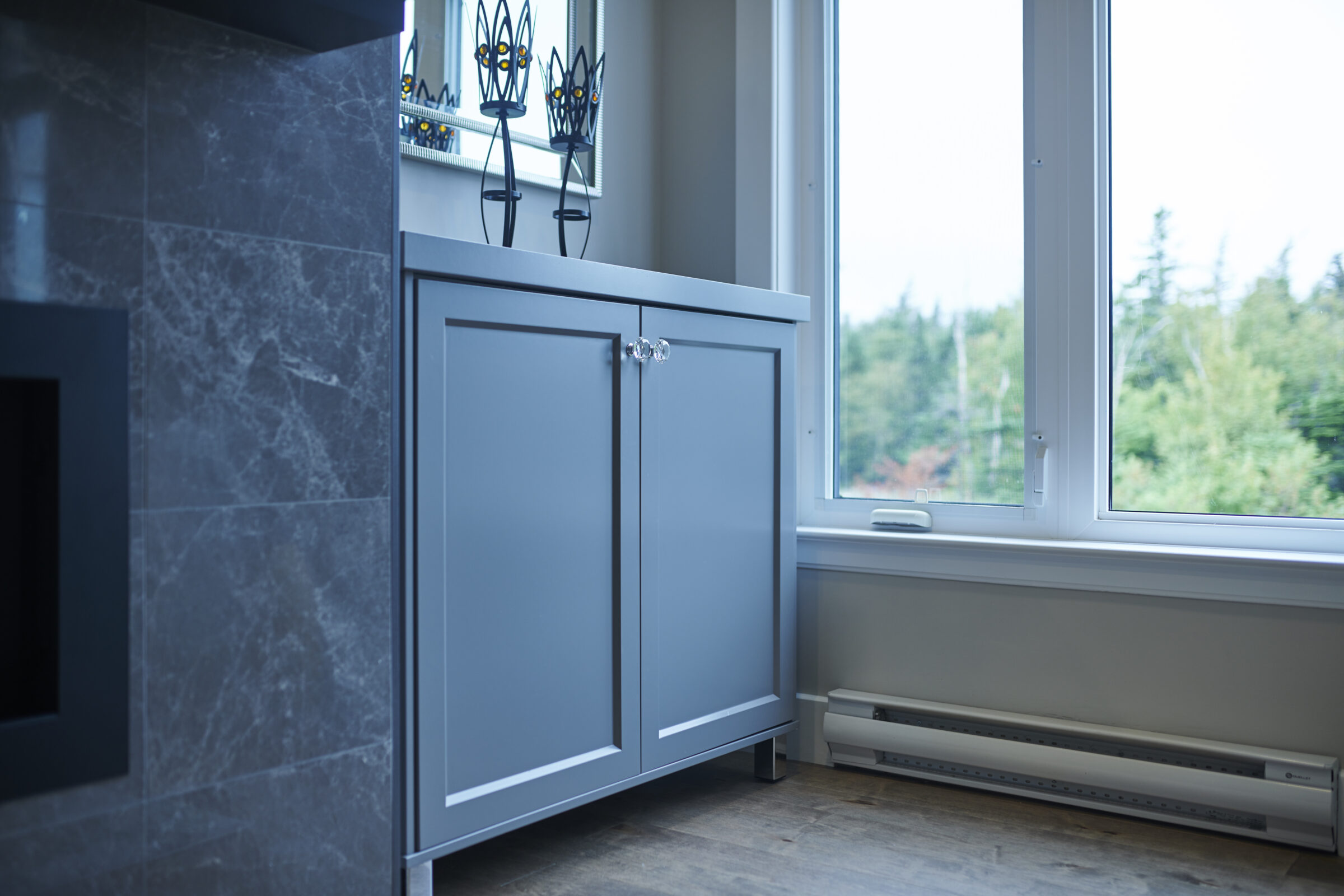 A modern interior corner with a blue-gray cabinet, marble wall tiles, decorative candle holders on top, beside a large window overlooking green trees.