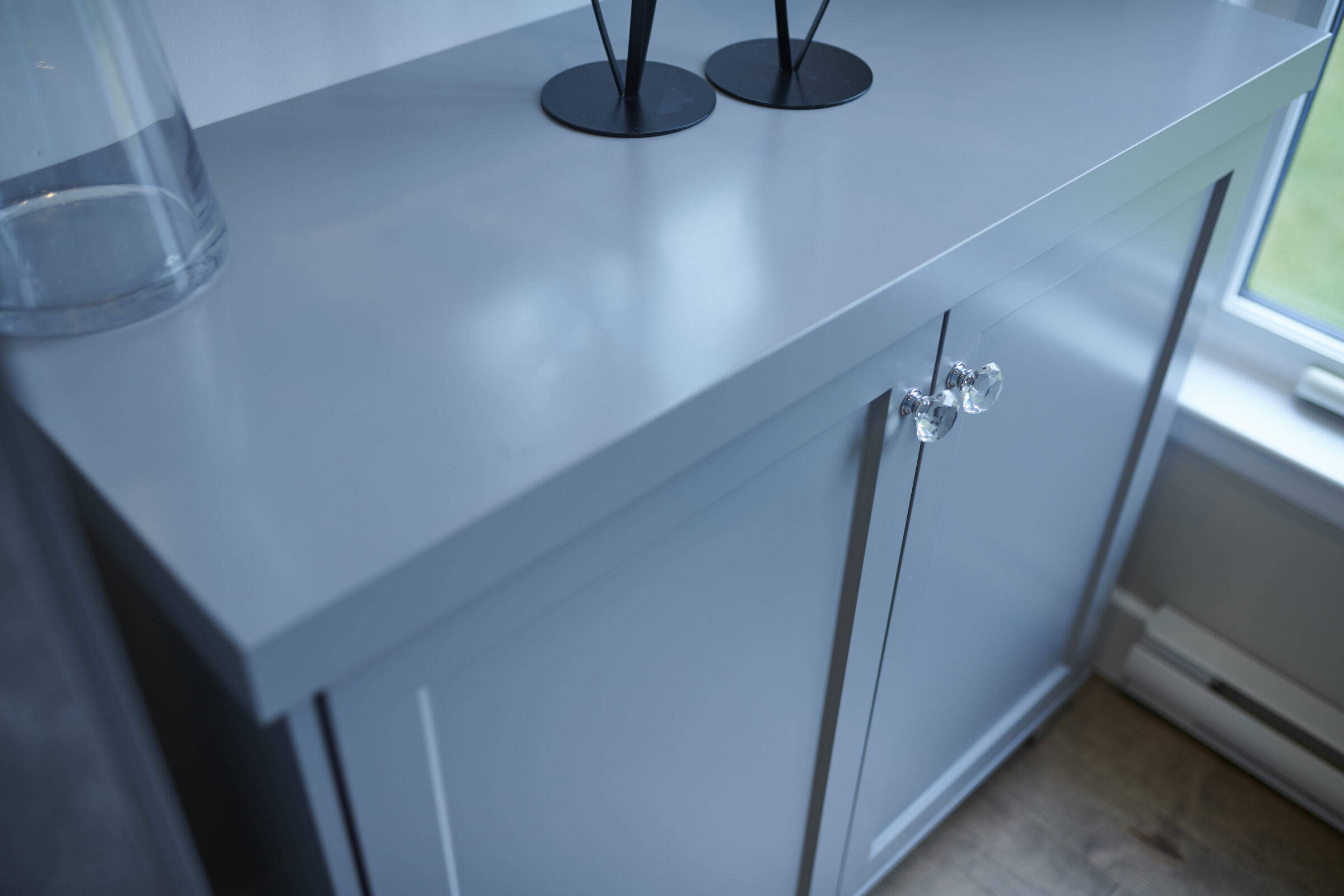 A modern, sleek grey cabinet with clear glass knobs. On the countertop, there’s a transparent vase and two black stands, beside a bright window.