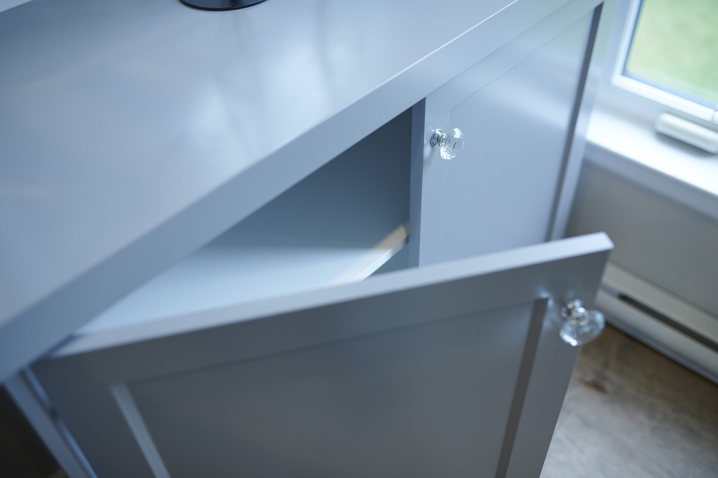 This image shows an open gray cabinet drawer with a clear knob, revealing an empty space inside, near a window with a view of greenery.