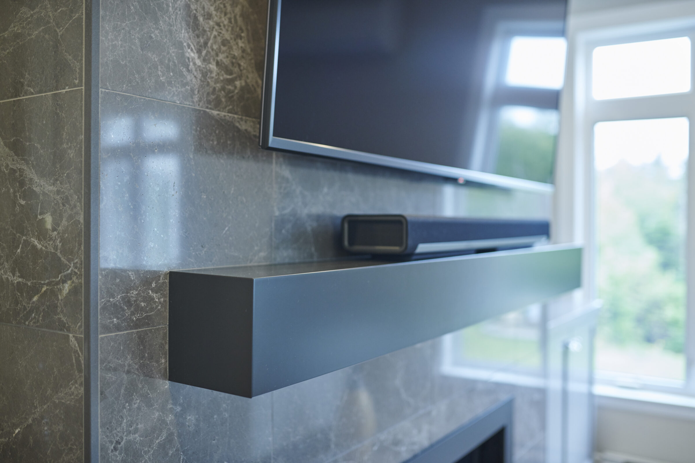 A flat-screen television is mounted on a marble wall above a modern, sleek fireplace. A soundbar rests on the narrow mantle, with windows letting in daylight.