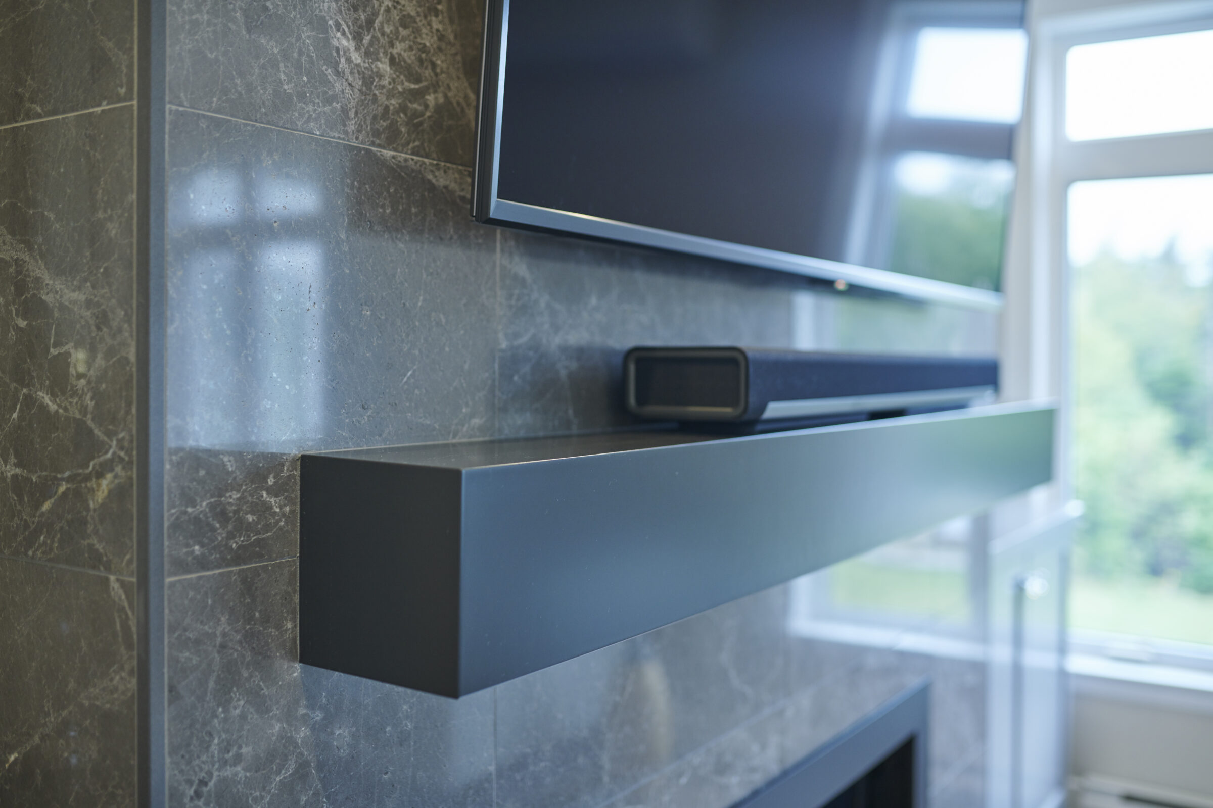 A modern living room with a wall-mounted television above a soundbar, both on a gray, marble-tiled wall, beside a window with a green view.