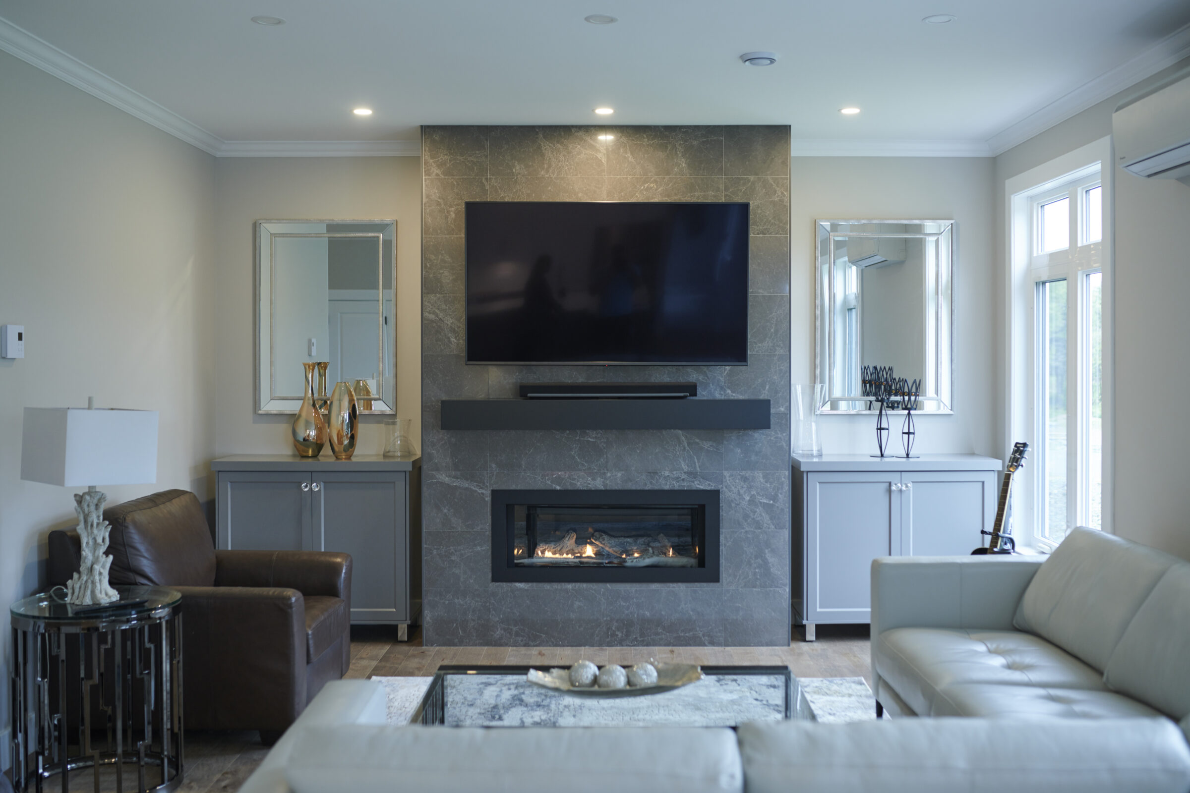 A modern living room featuring a marble fireplace, mounted flat-screen TV, cozy furniture, decorative mirrors, and a well-lit interior space.