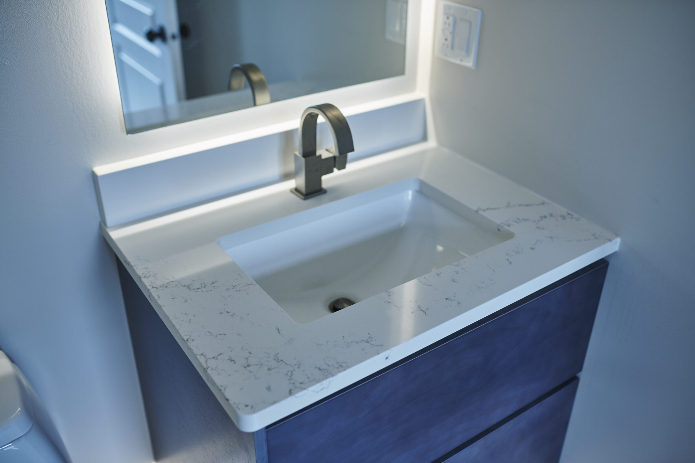 Modern bathroom with under-lit mirror above marble countertop, integrated sink, single faucet, blue vanity cabinet, and wall-mounted switches. Clean, minimalist design.