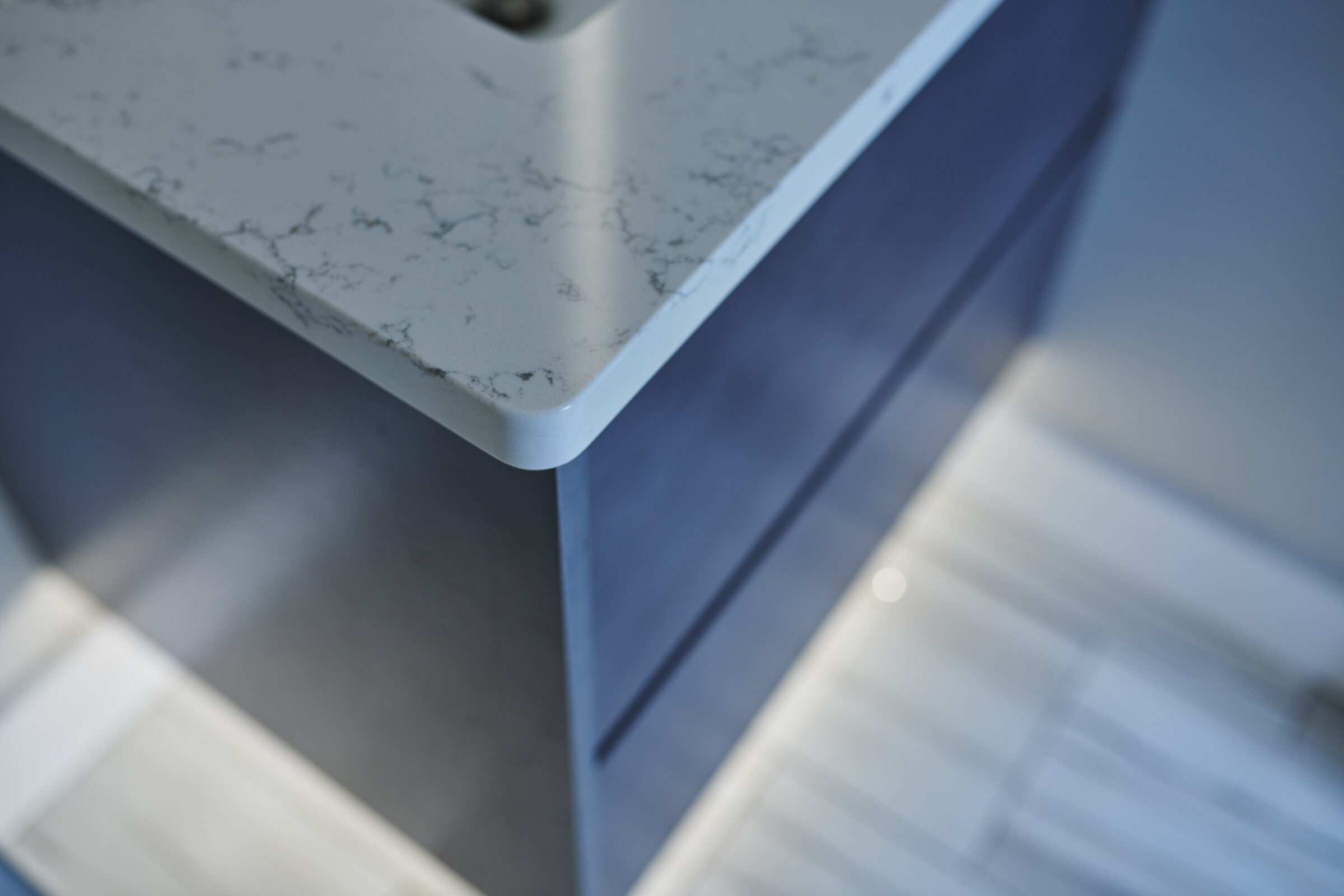 This image shows a close-up corner of a marble countertop with a blurred blue cabinet beneath it, reflecting a modern and sleek interior design.