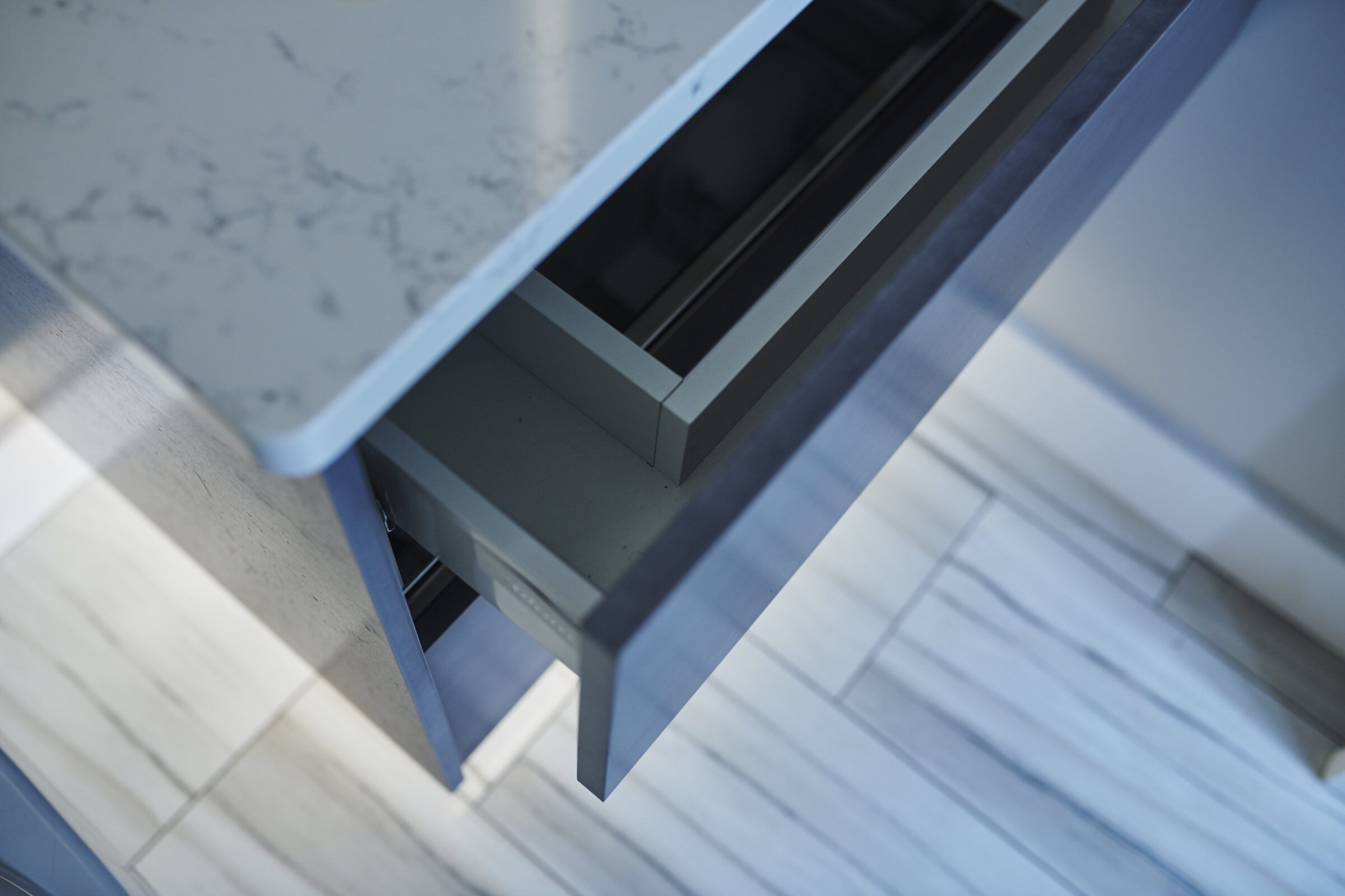 The image shows a close-up angle of an open, gray, modern kitchen drawer, with a marble countertop above, and tiled flooring below.
