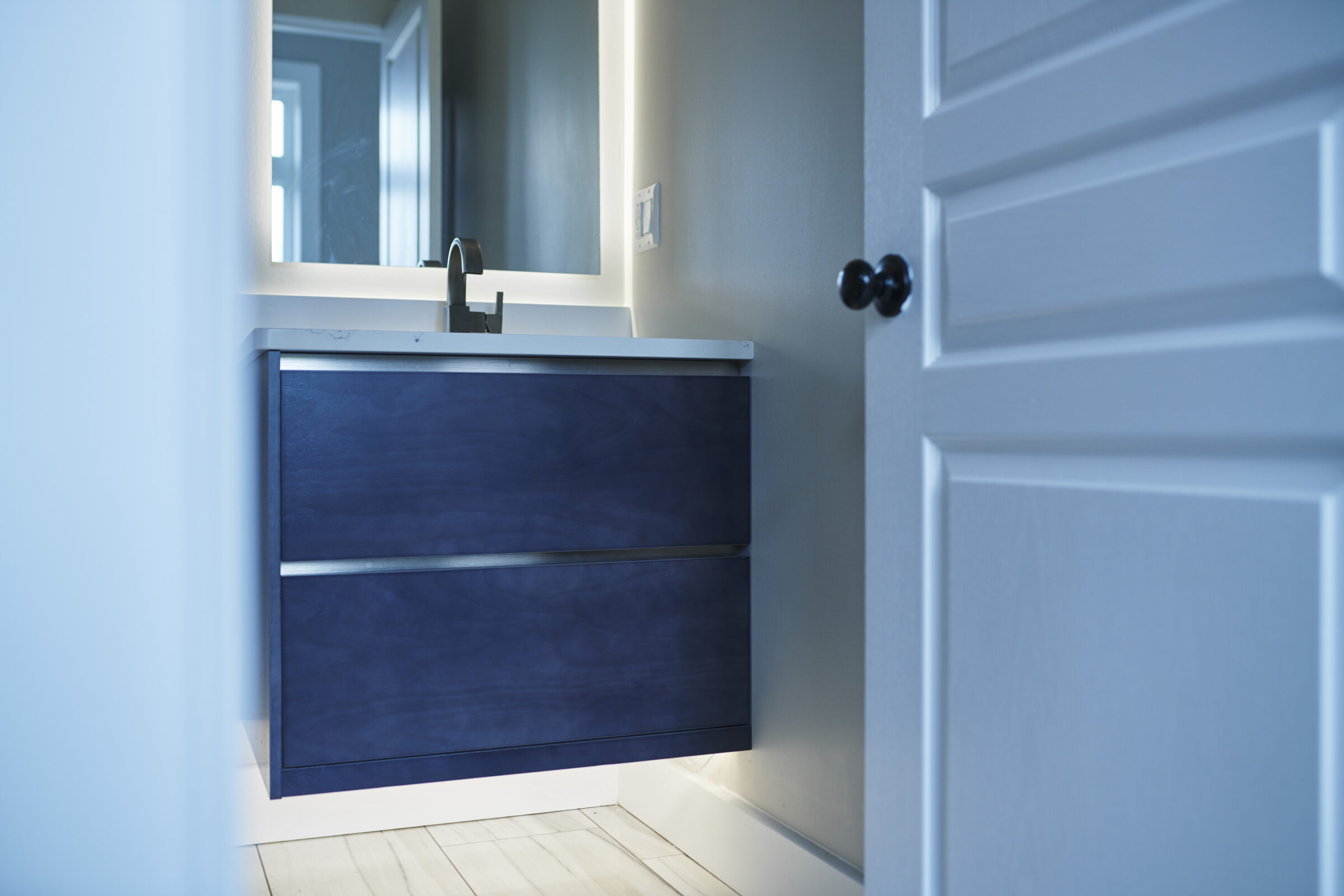 A modern bathroom vanity with a dark blue cabinet, white countertop, and black faucet is partially visible through an open door.