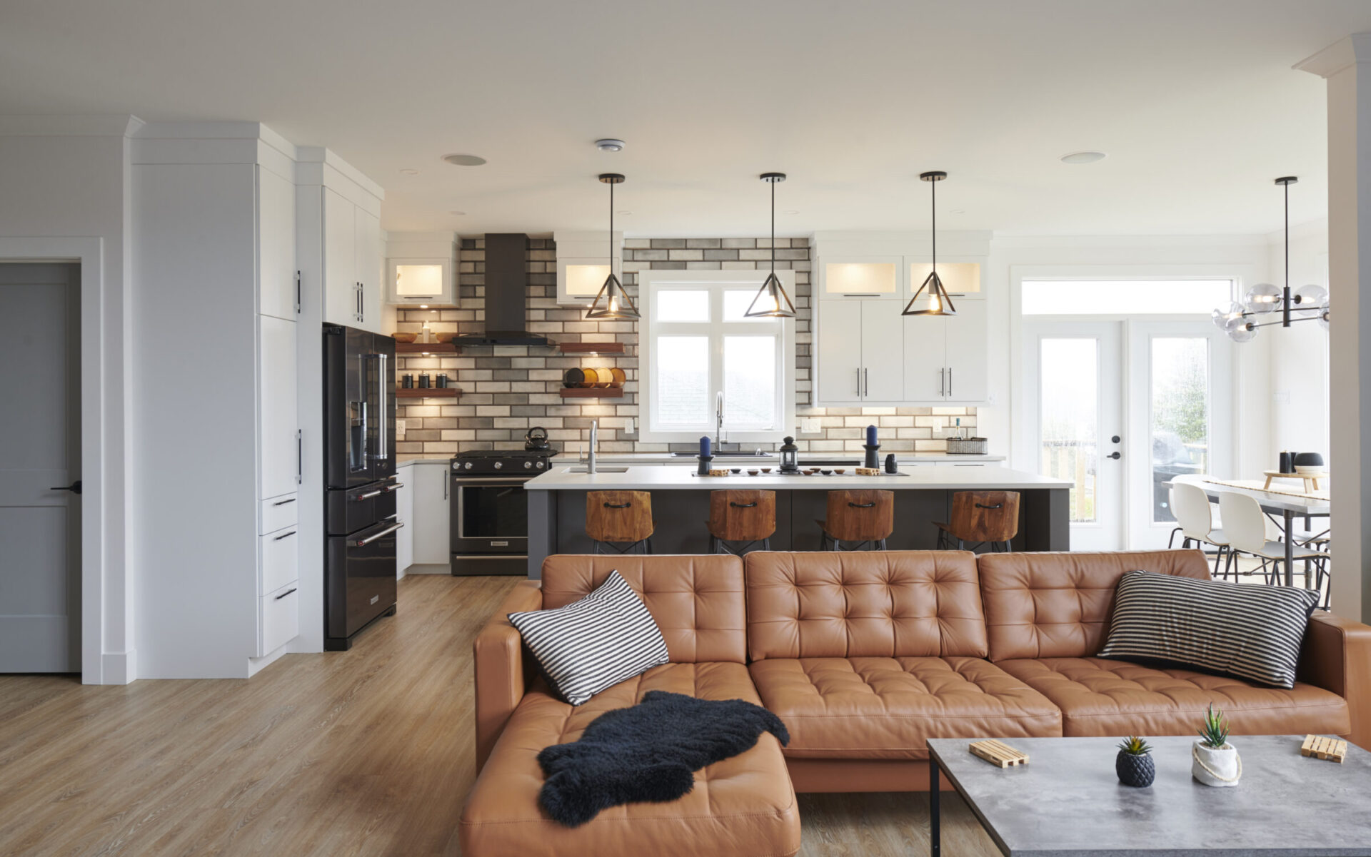 Modern open-plan living space with a kitchen, dining area, and living room. Brown leather couch, wooden stools, elegant pendant lights, and neutral tones.