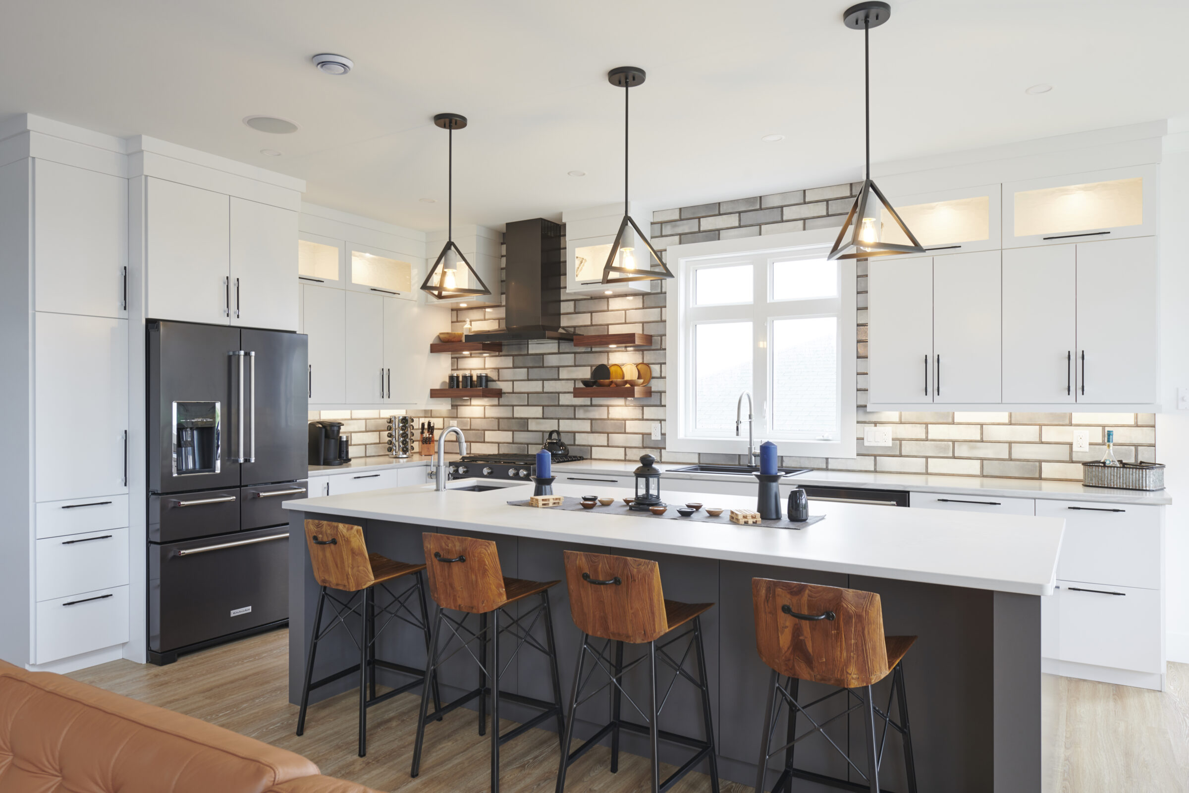 This is a modern kitchen interior with white cabinets, stainless steel appliances, bar stools, pendant lights, and a brick-style backsplash.