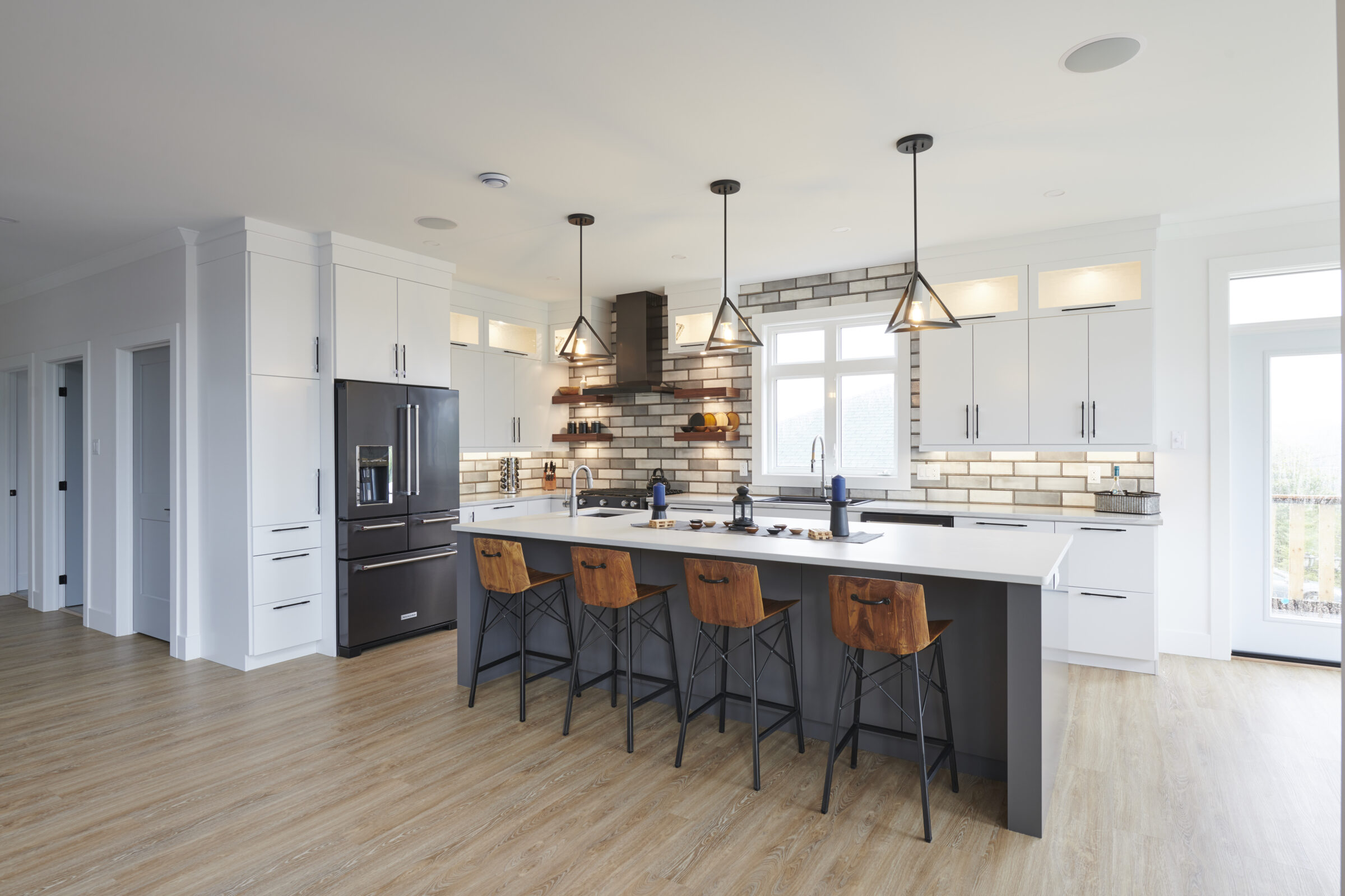 Modern kitchen with white cabinets, black appliances, wood floor, central island, pendant lights, and wooden stools. Bright, clean, and contemporary style.