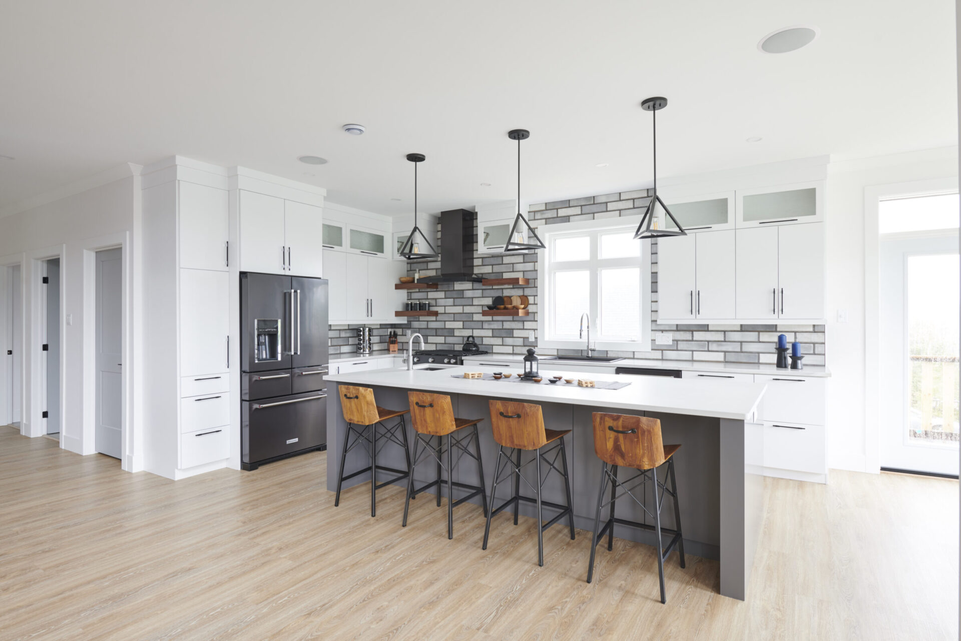 Modern kitchen with white cabinetry, gray backsplash, black appliances, a central island with bar stools, pendant lights, and light wood flooring.