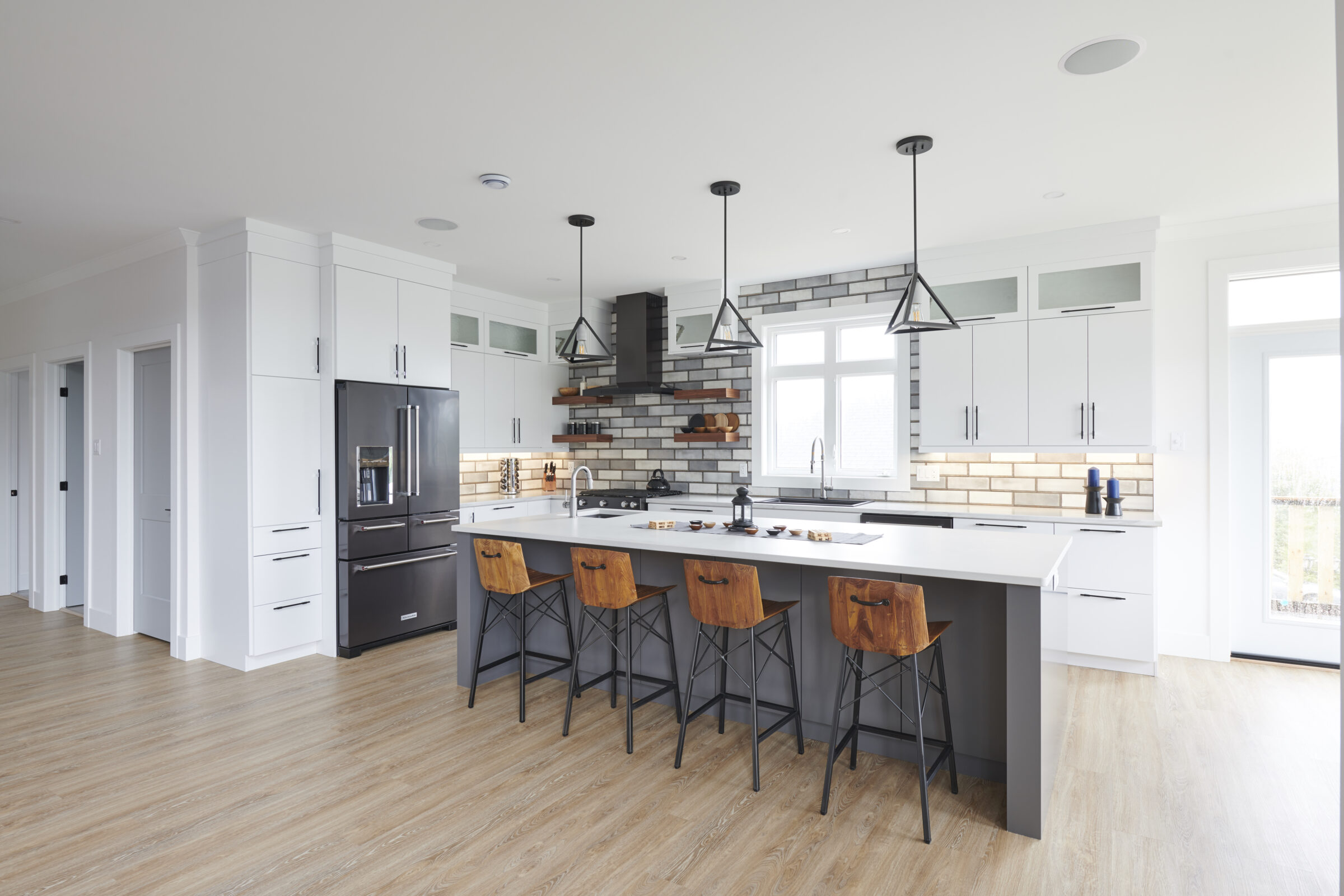 Modern kitchen with black appliances, white cabinetry, wooden floor, a central island with stools, pendant lights, and a brick backsplash.