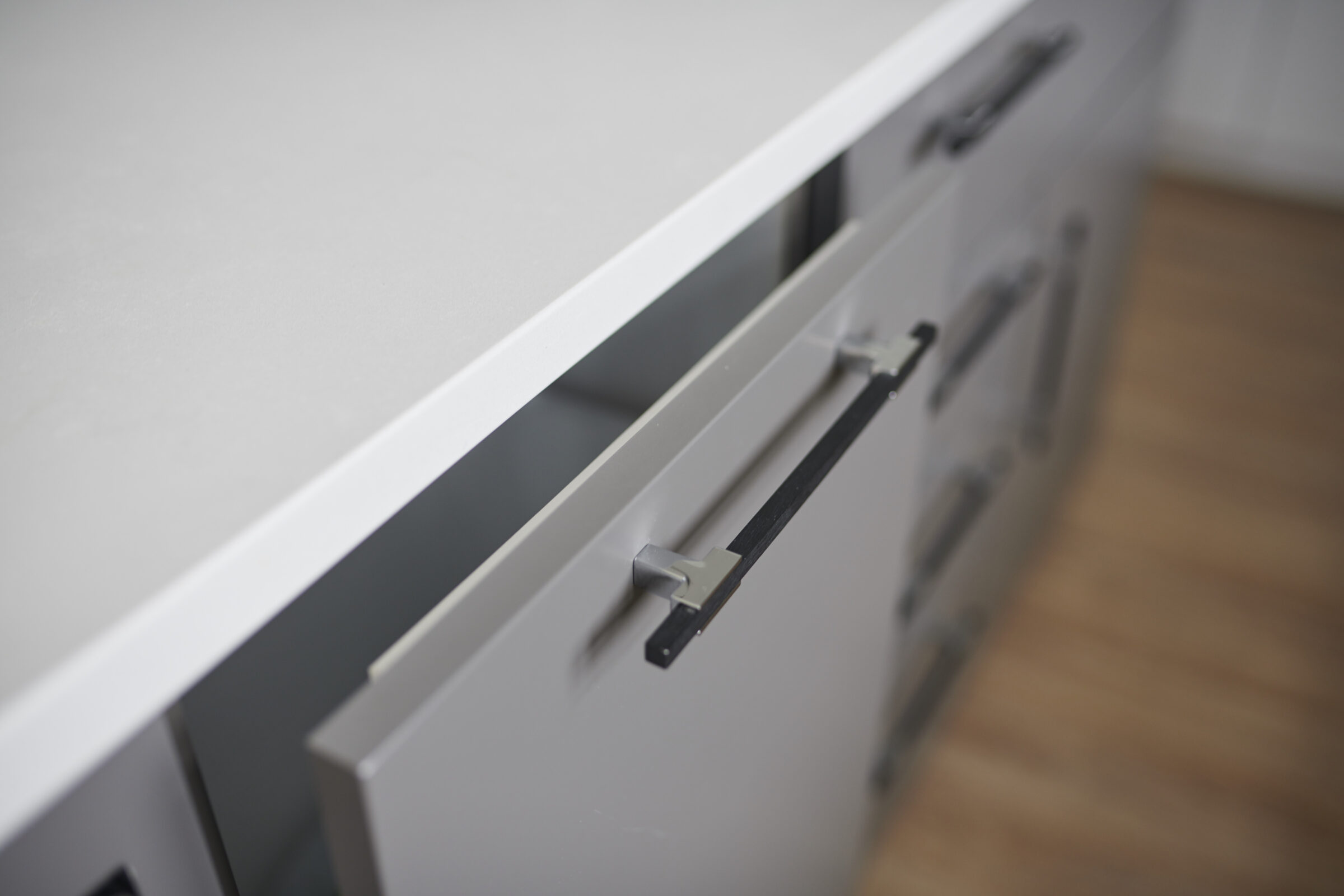 A modern kitchen cabinet with a slightly open drawer, showcasing its minimalistic design and sleek handle against a wooden floor background.