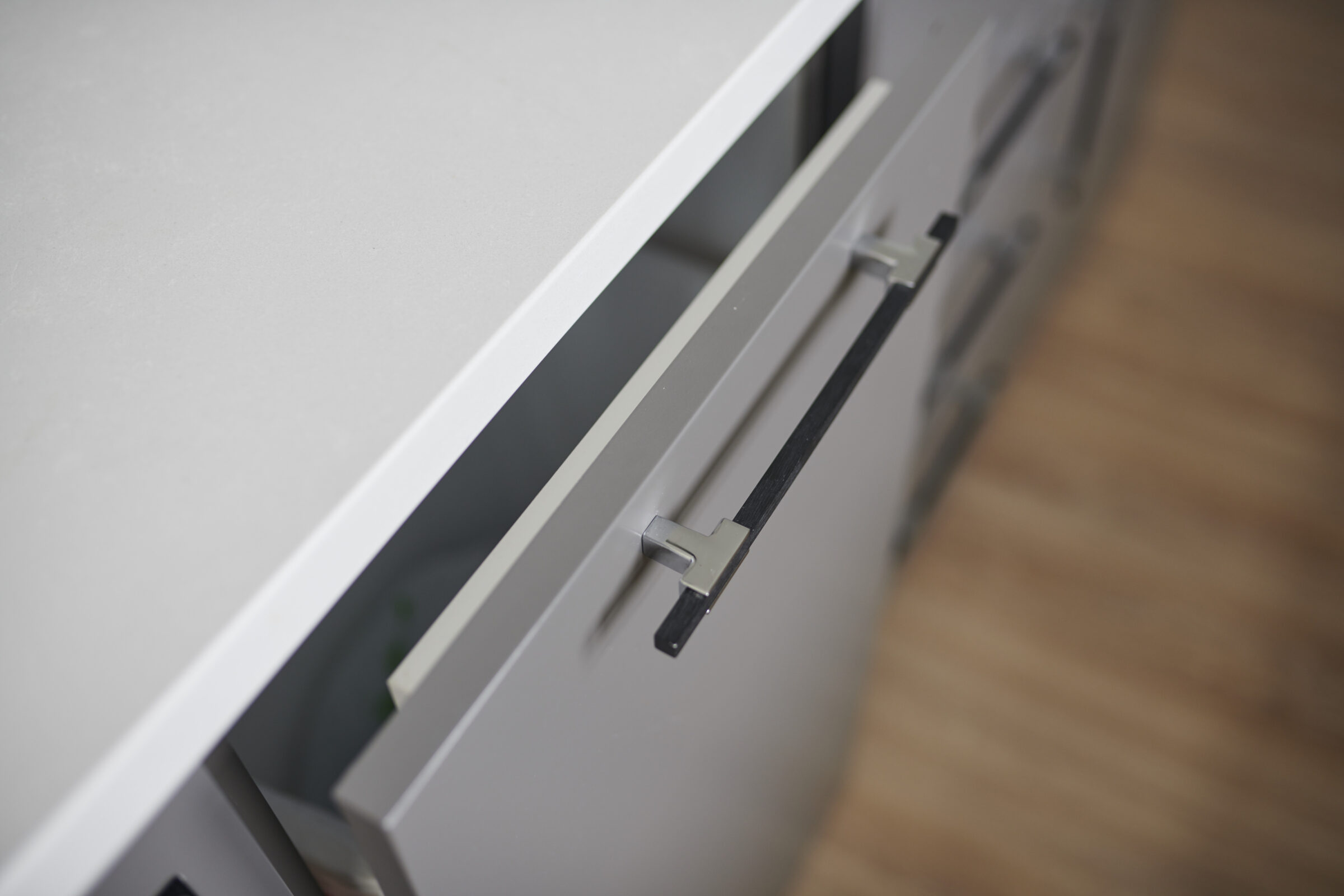 The image shows a close-up of an open drawer with minimalistic metal handles, set against a gray cabinet and a wooden floor background.