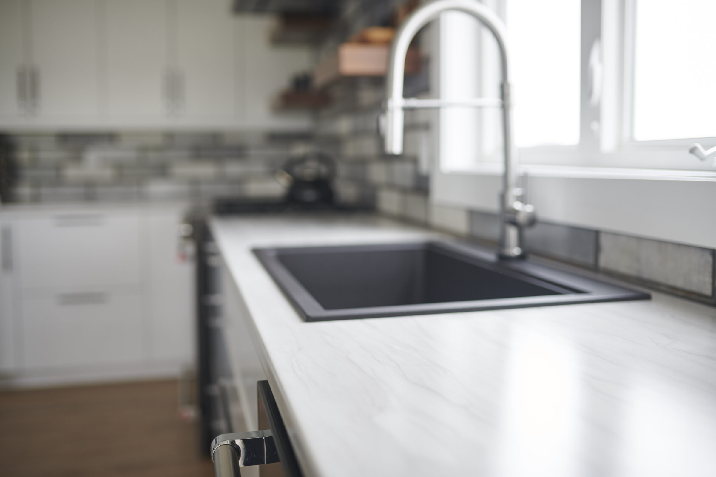 Modern kitchen interior with a blurred background. Features white marble countertop, black sink, and a stylish faucet near a bright window.