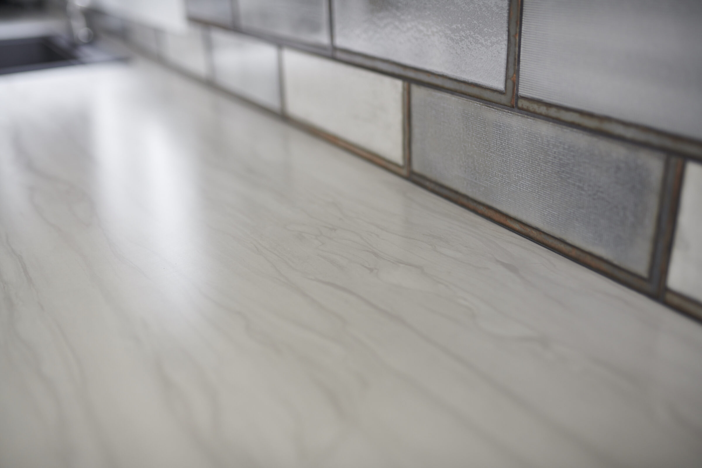 The image shows a close-up of a marble countertop with reflective metal border tiles on a kitchen backsplash, under soft, diffuse lighting.