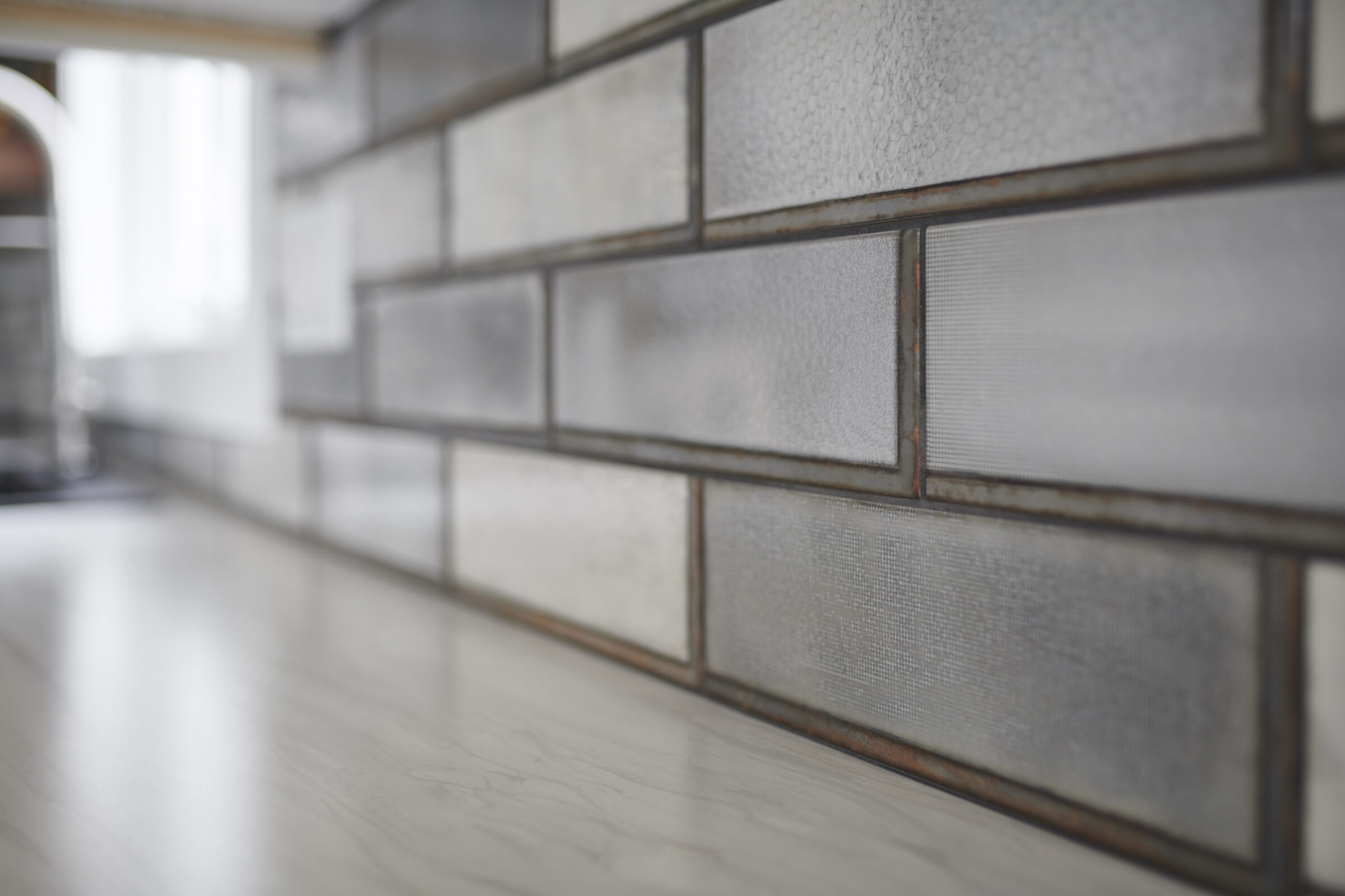 This image shows a close-up view of a textured tiled backsplash in a kitchen, with a blurry background, highlighting the tiles' intricate details.