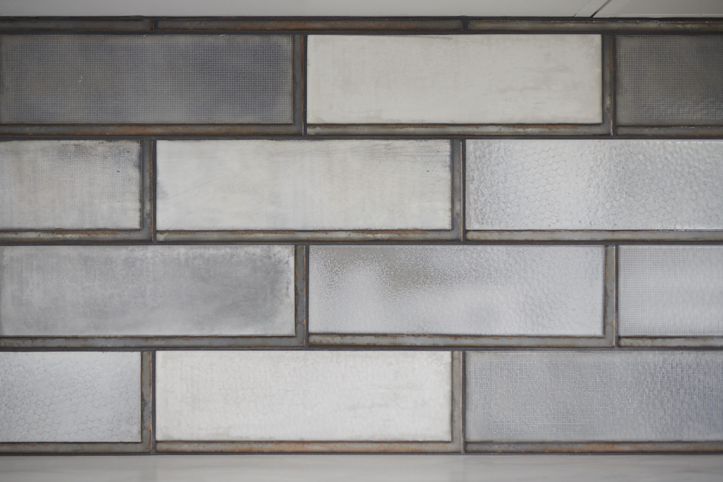 The image shows a wall with varied textured glass tiles framed in dark grout, offering a mix of translucency and opacity with a modern aesthetic.