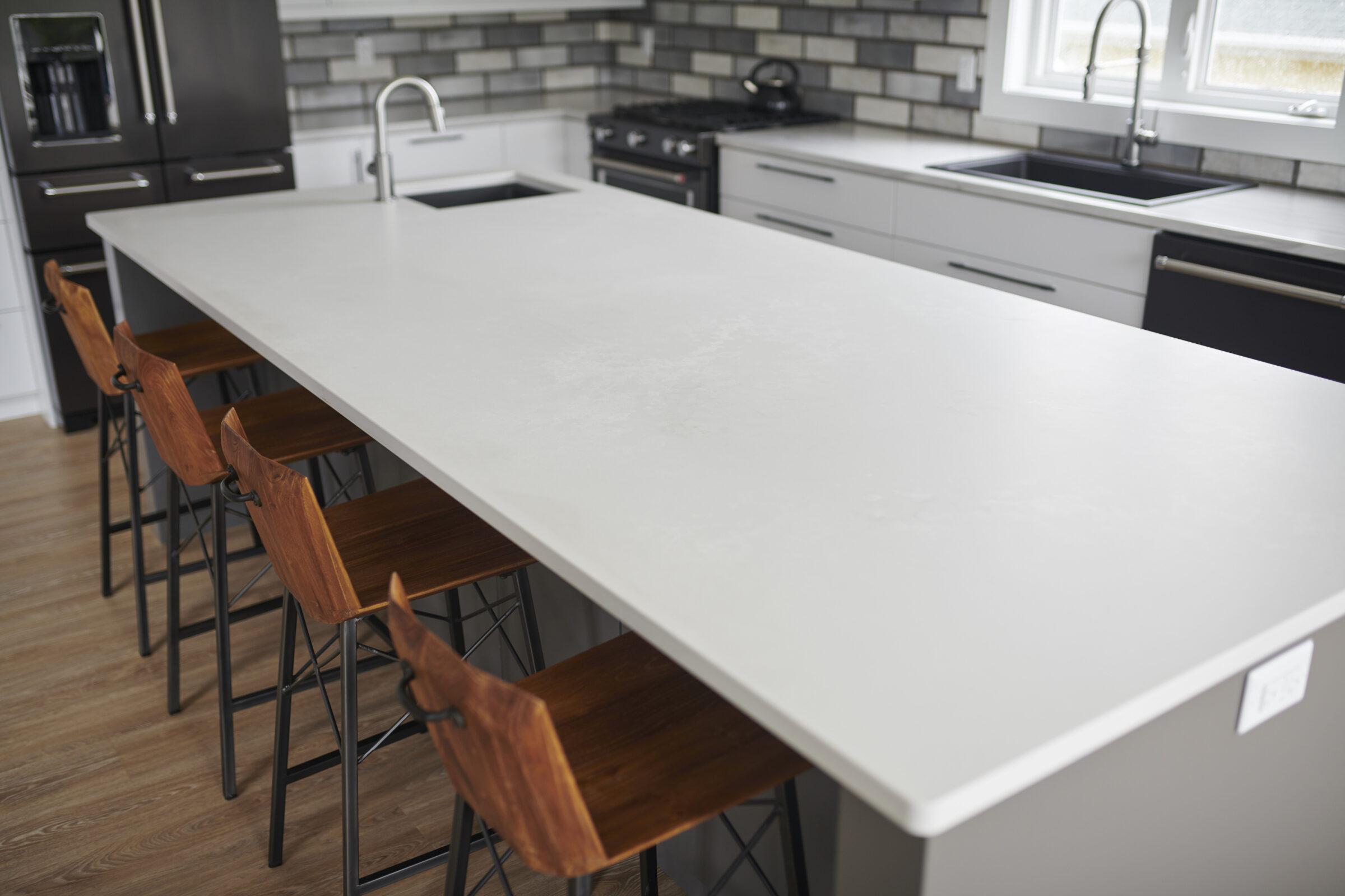 A modern kitchen interior with a large white island countertop and wooden stools. Black appliances and gray tile backsplash are visible.