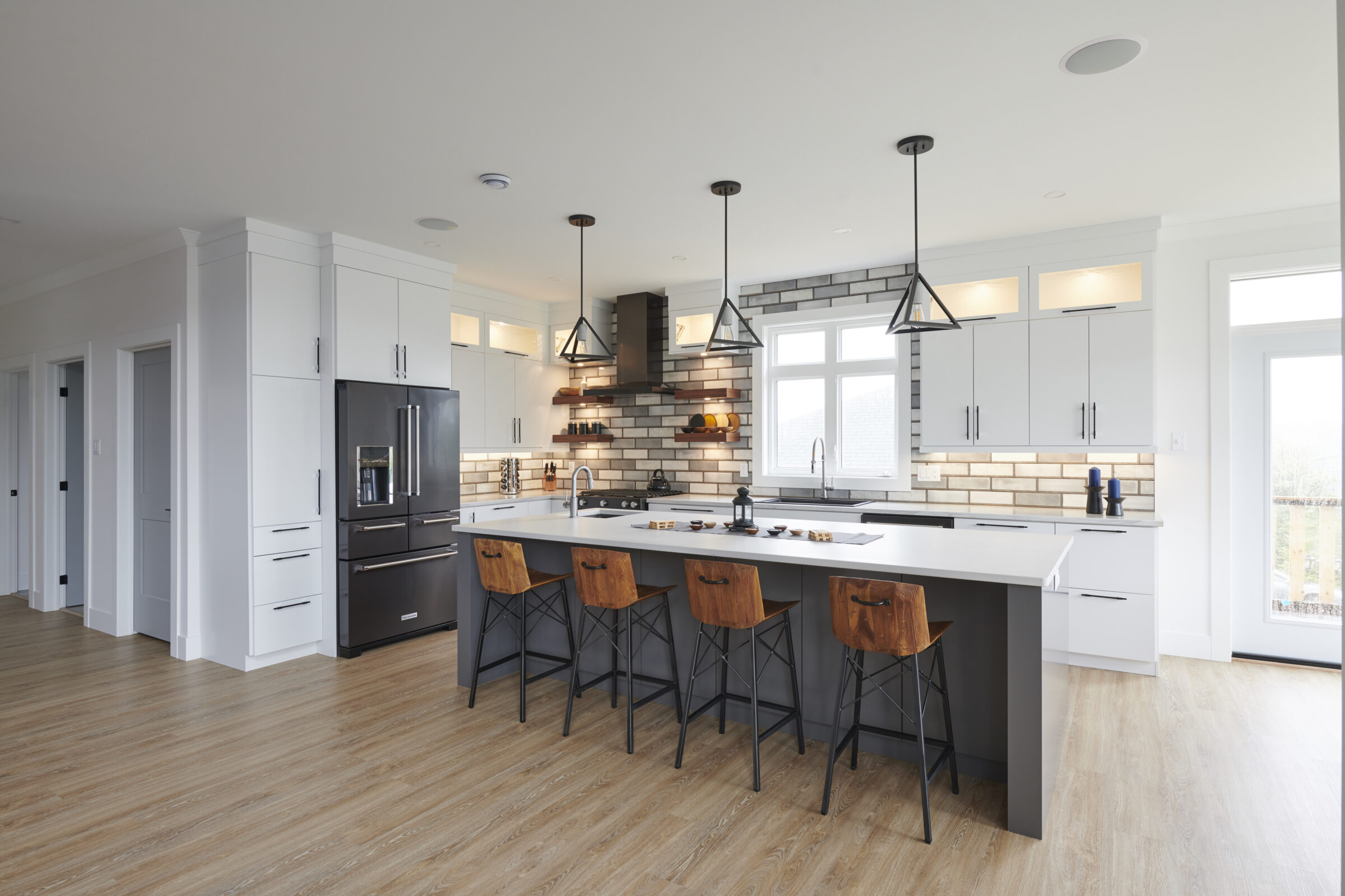 A spacious modern kitchen with white cabinets, black appliances, wooden floors, and a central island with bar stools under pendant lights.