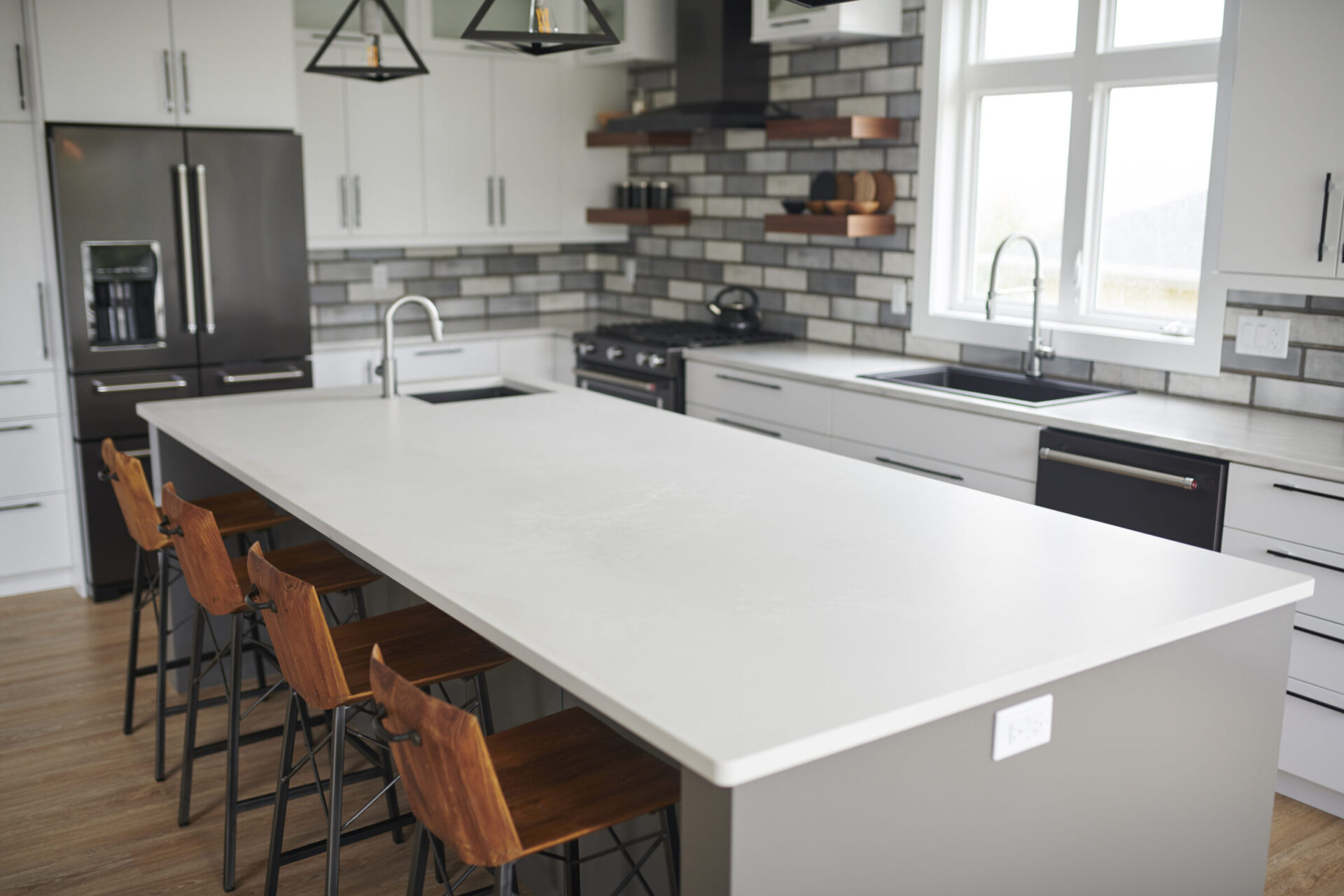 A modern kitchen with a large island, wooden chairs, stainless steel appliances, white countertops, gray cabinets, and a subway tile backsplash.