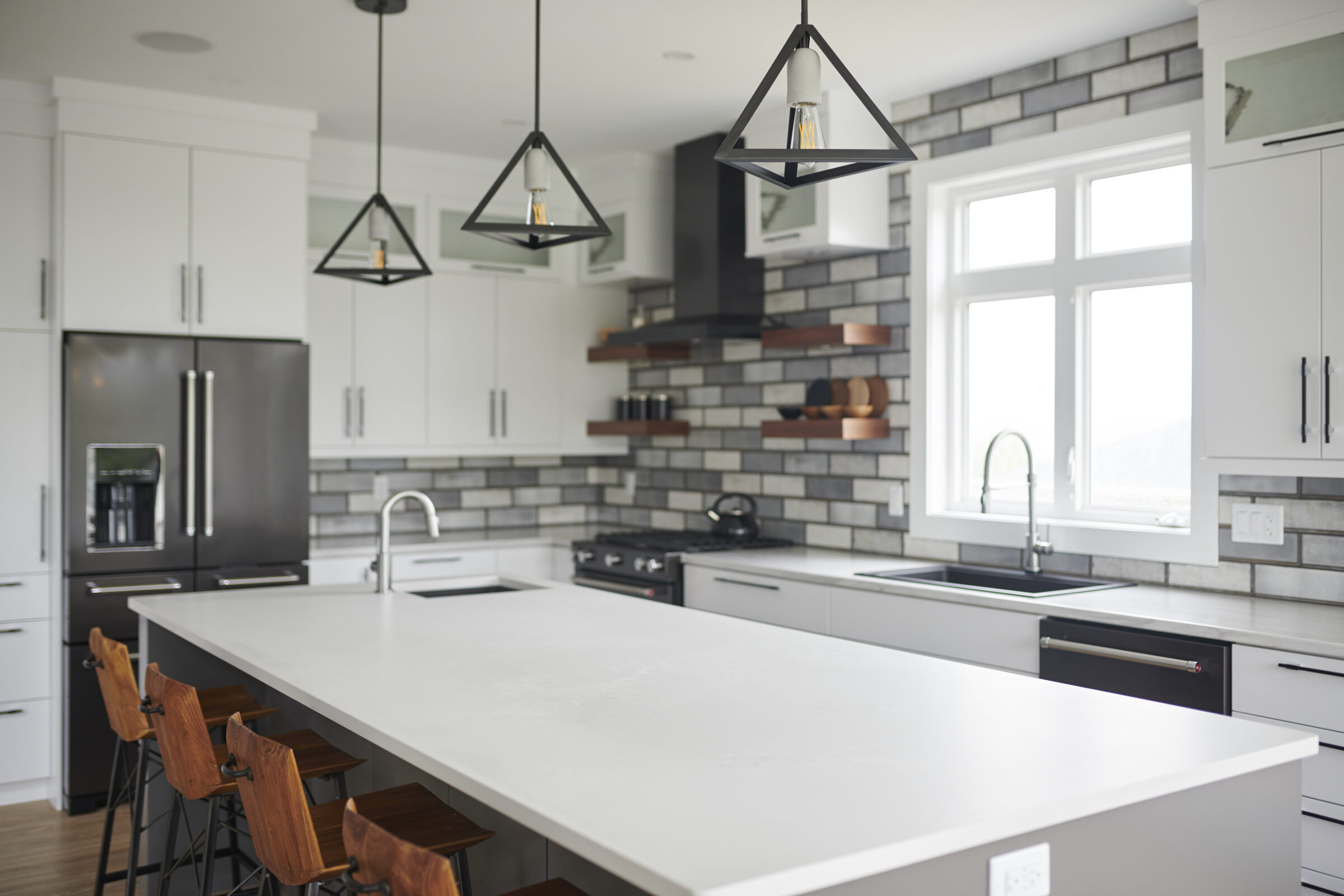 Modern kitchen interior with a large island, wooden stools, white cabinets, stainless steel appliances, and pendant lights above the counter.
