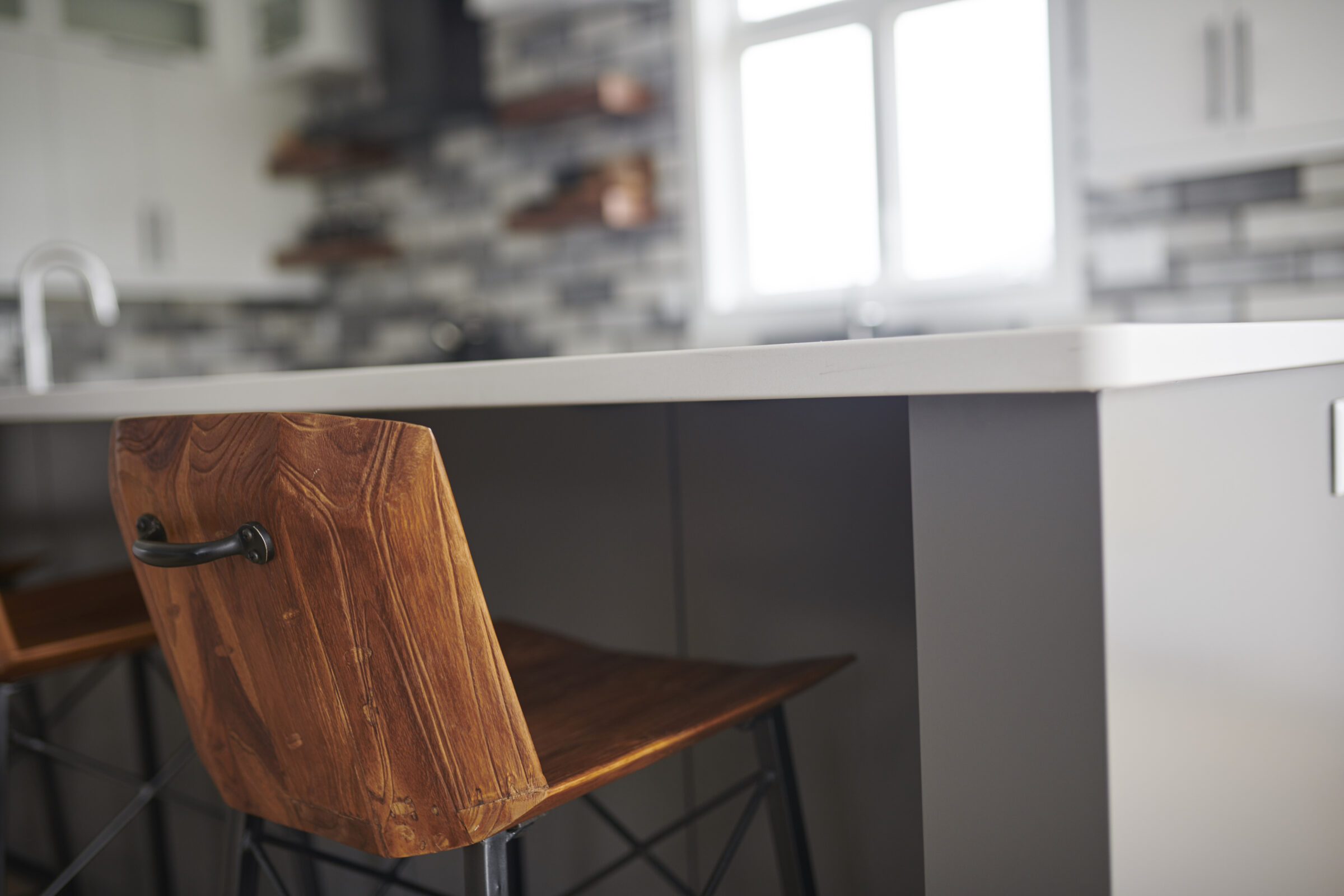 A modern kitchen featuring white countertops, grey cabinets, and a wooden bar stool. There's a tiled backsplash and natural light from windows.
