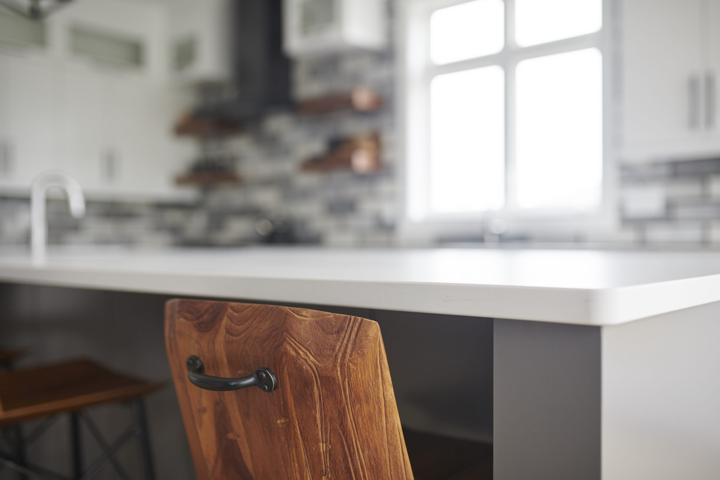 Modern kitchen interior focused on a wooden chair with a black handle, white countertop, tiled backsplash, and natural light from windows.