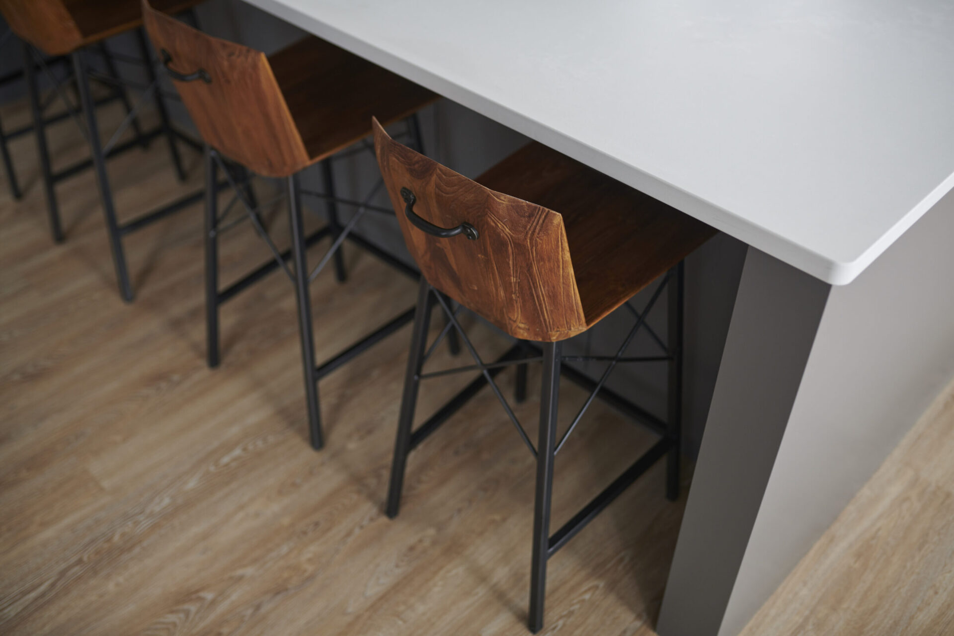 A modern kitchen interior with a white countertop, wooden bar stools with black metal legs, and light wooden flooring, shot in a minimalist style.