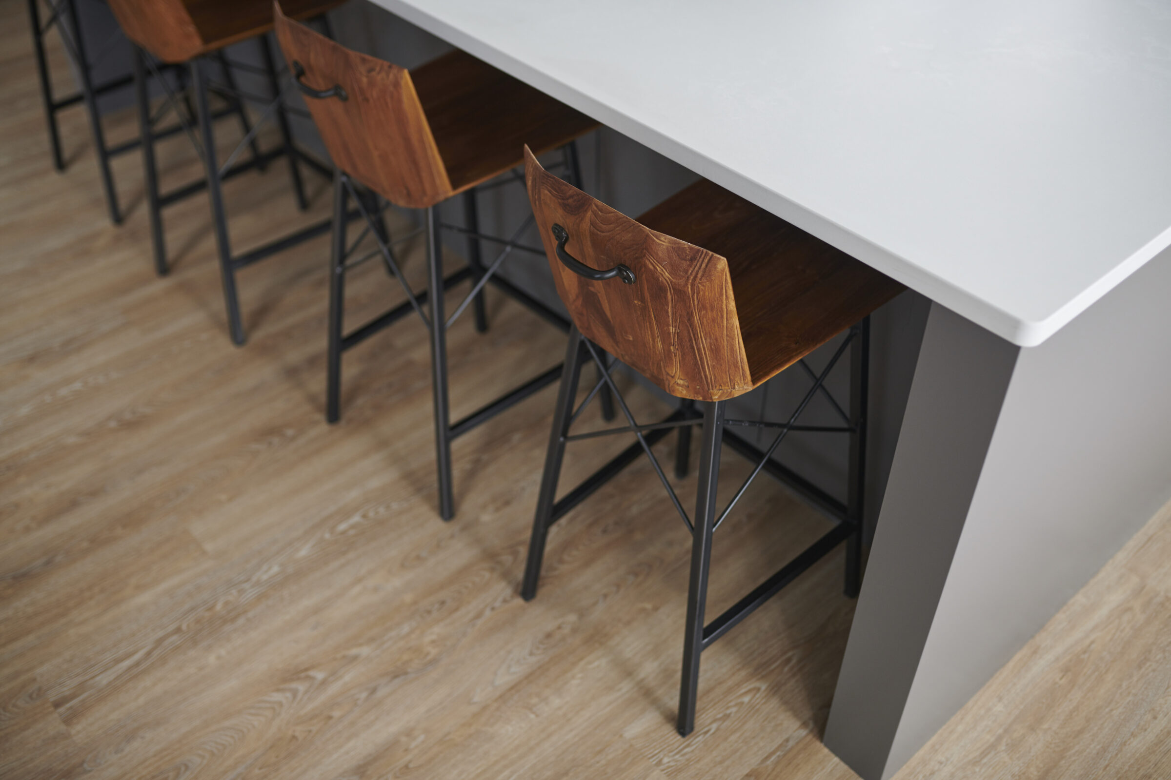 A modern kitchen with a white countertop and stylish wooden stools having black metal legs arranged on a wooden herringbone-patterned floor.