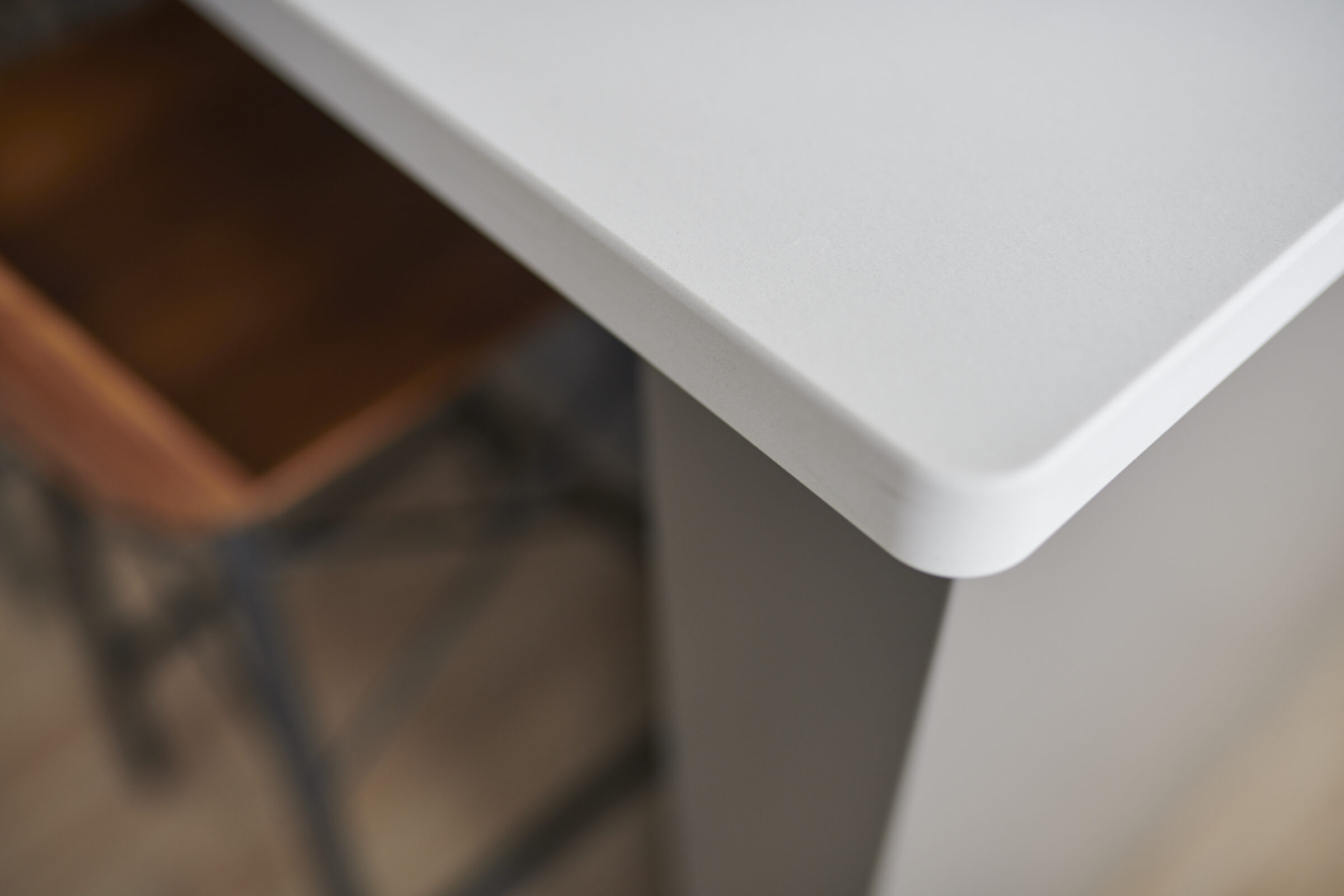 This image shows a close-up of a white table corner with a blurred wooden chair and metal frame in the background, illustrating modern interior design.