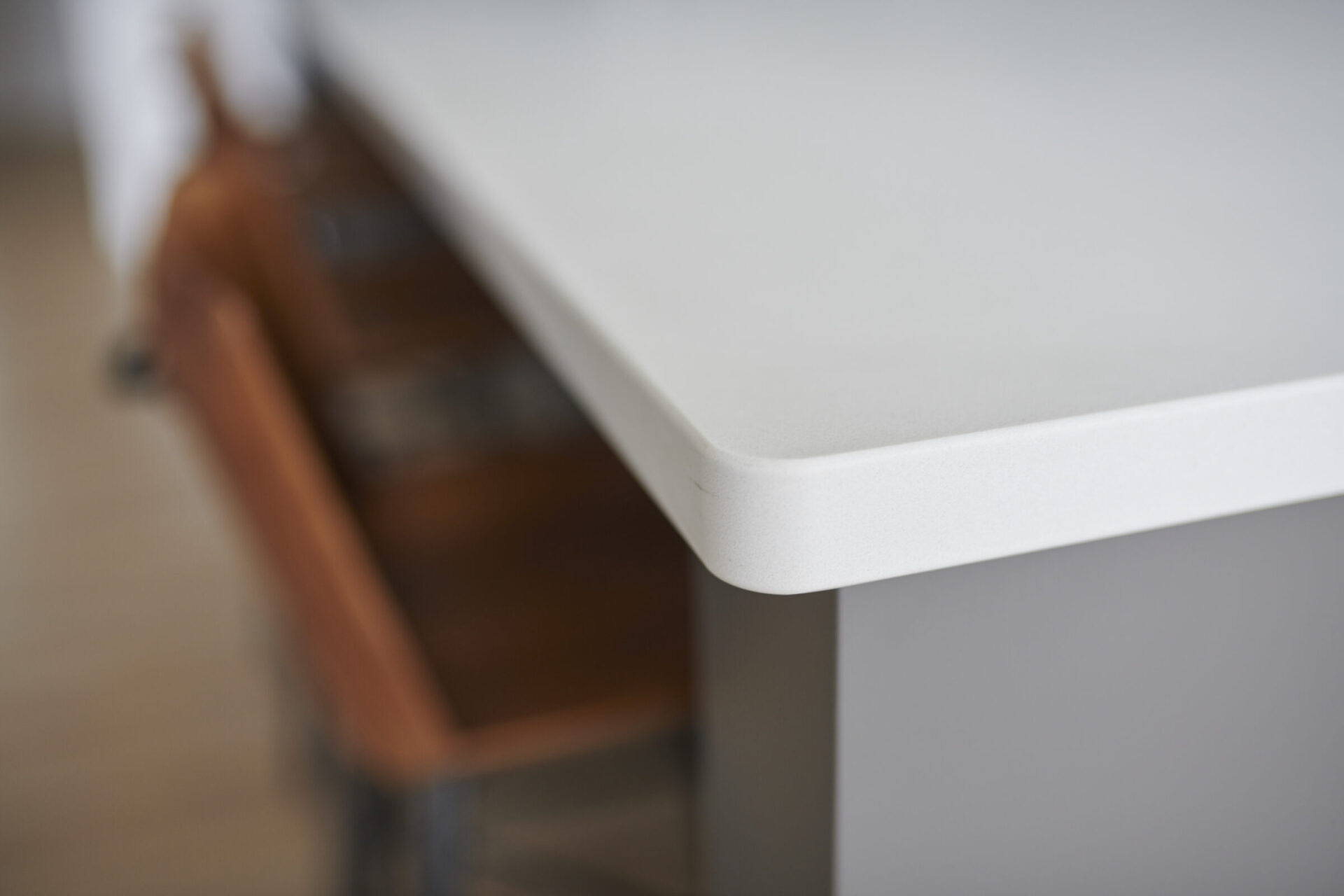 A close-up photo highlighting the edge of a white table with a blurred wooden chair in the background, emphasizing texture and simple modern design.