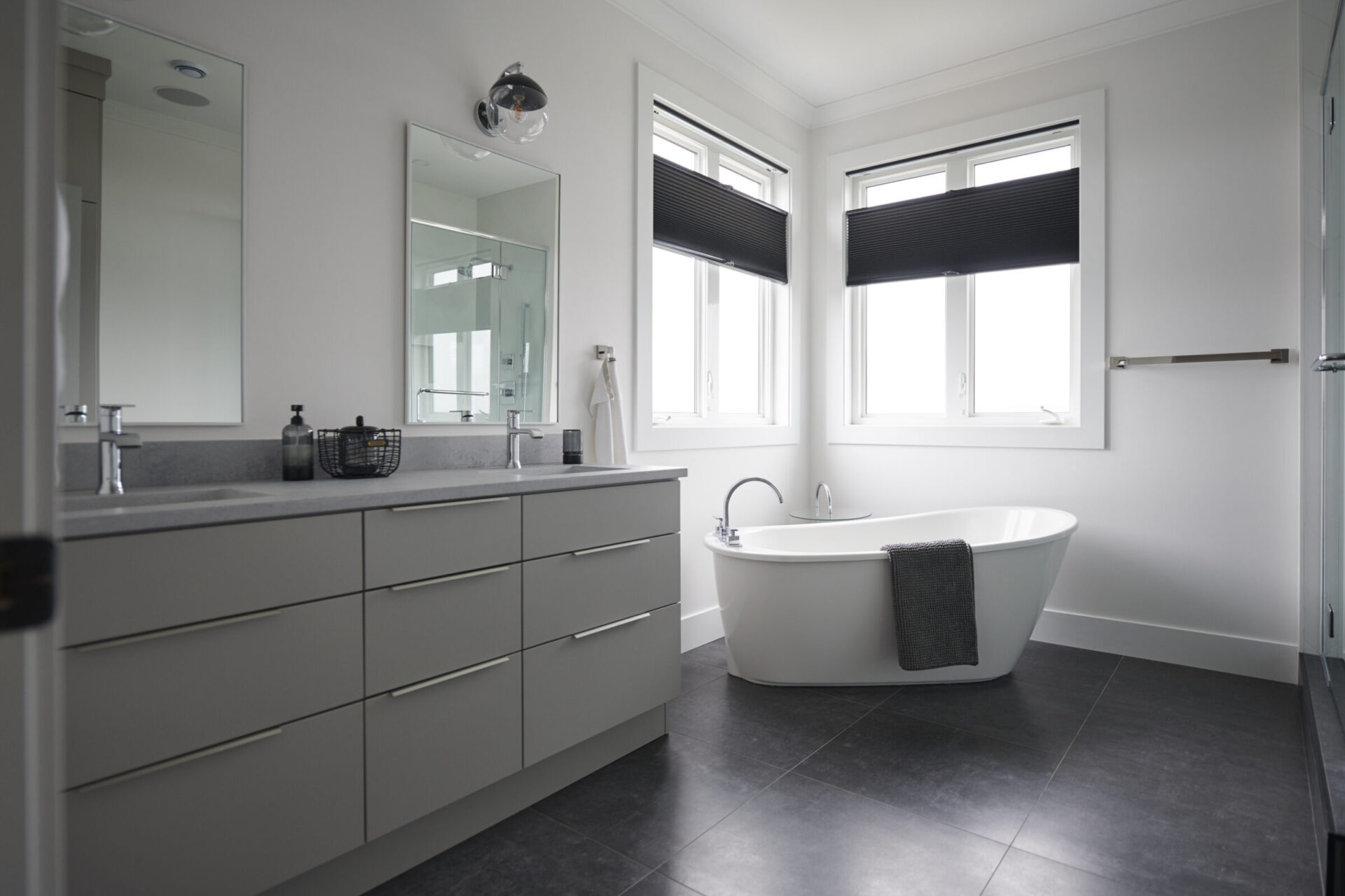 This is a modern, clean bathroom with a large mirror, a double sink vanity, dark floor tiles, a freestanding tub, and black window blinds.