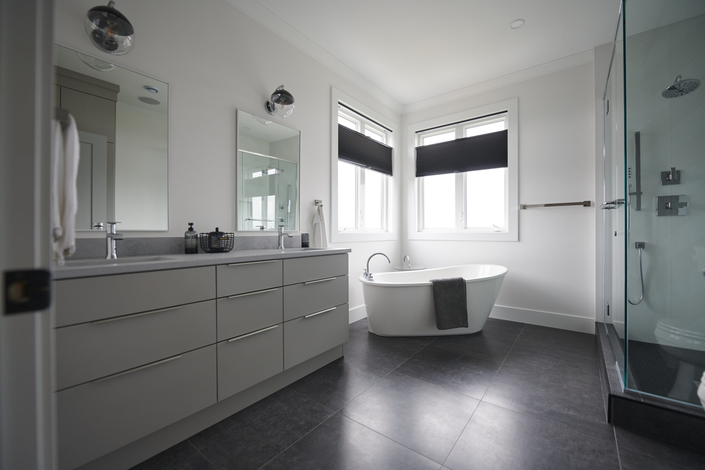 Modern bathroom interior with twin sinks, vanity, freestanding tub, glass shower enclosure, tiled floor, and windows with dark blinds. Clean and spacious design.