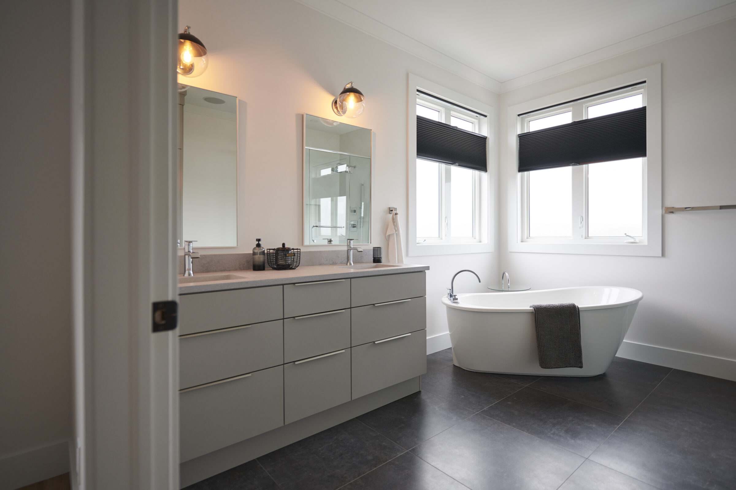 A modern bathroom with a standalone bathtub, double vanity with mirrors, dark floor tiles, and windows with black blinds. The setting is clean and bright.