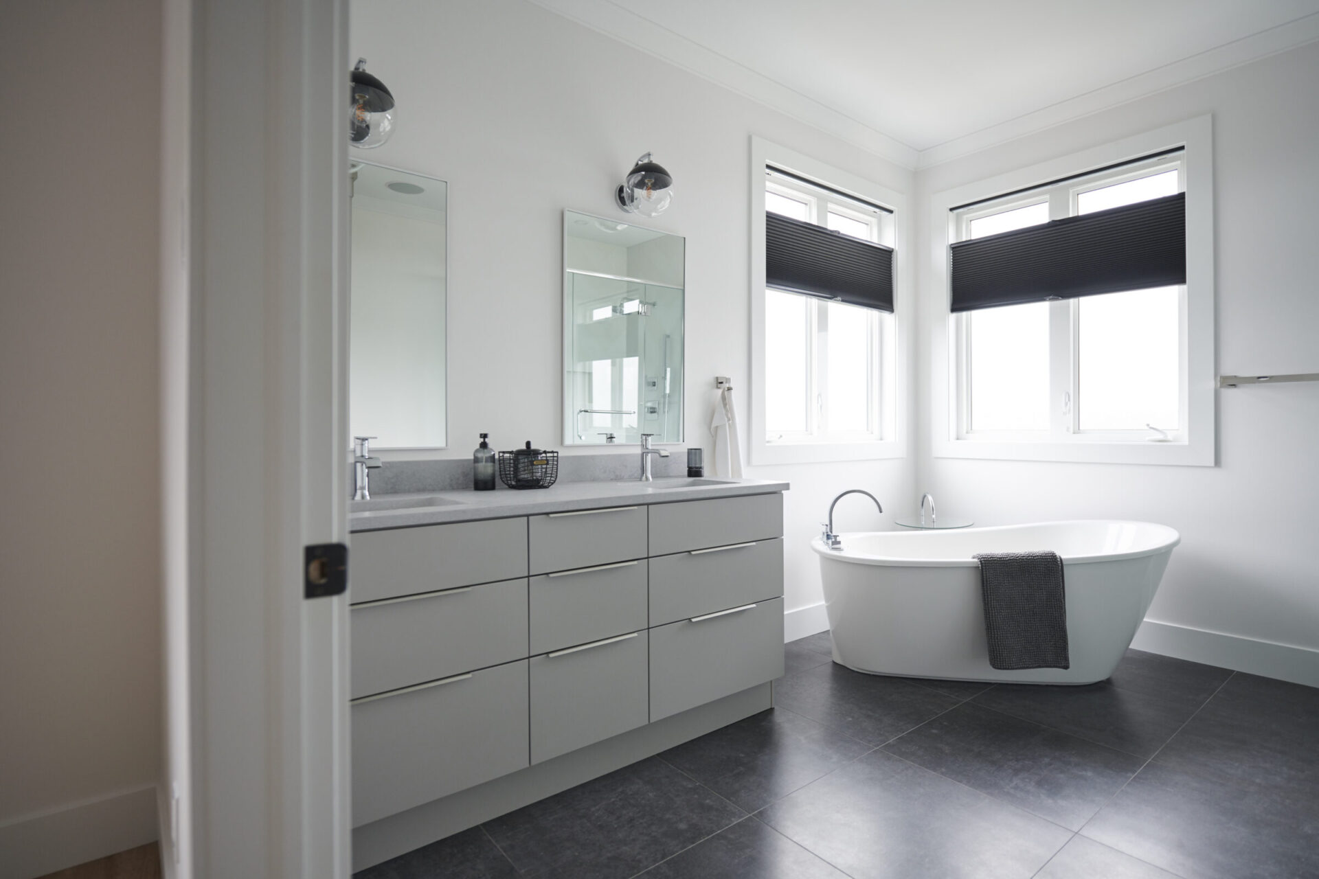This is a modern bathroom featuring a gray vanity with sink, a large mirror, a freestanding bathtub, dark tiled flooring, and blinds on the windows.