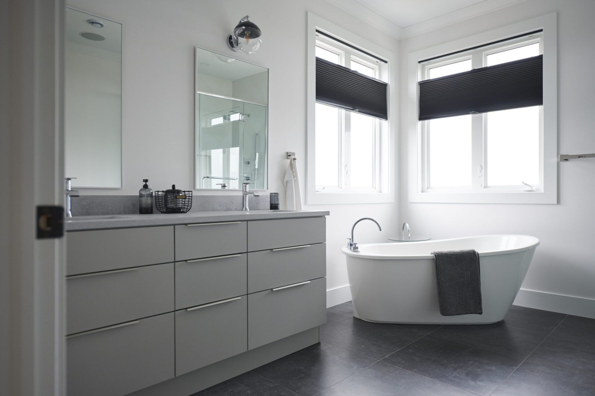 Modern bathroom interior with a large freestanding tub, double sink vanity, mirrors, and dark floor tiles. Natural light comes through two windows with blinds.