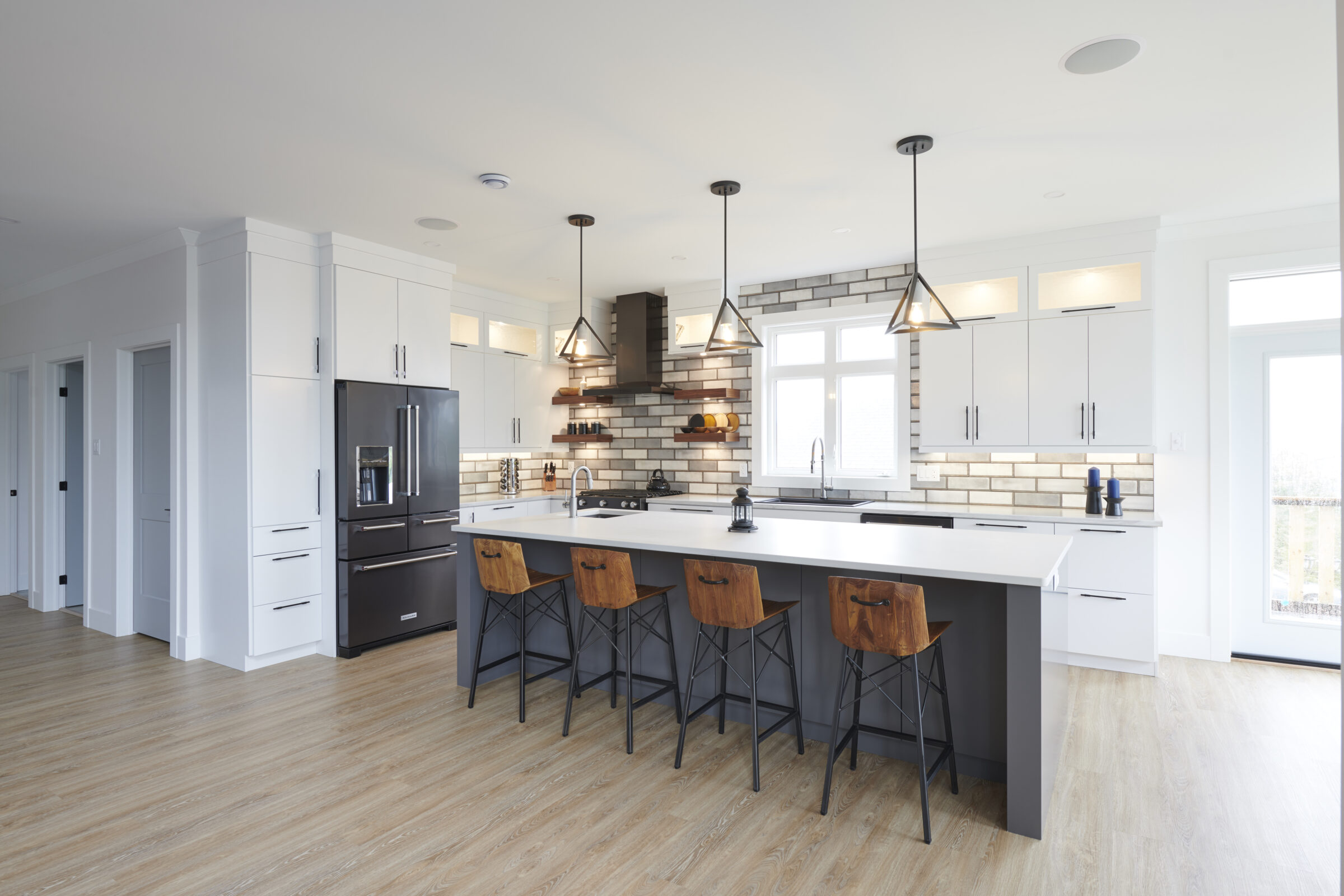 Modern kitchen interior with large island, bar stools, white cabinetry, stainless steel appliances, pendant lights, and a blend of natural and artificial lighting.