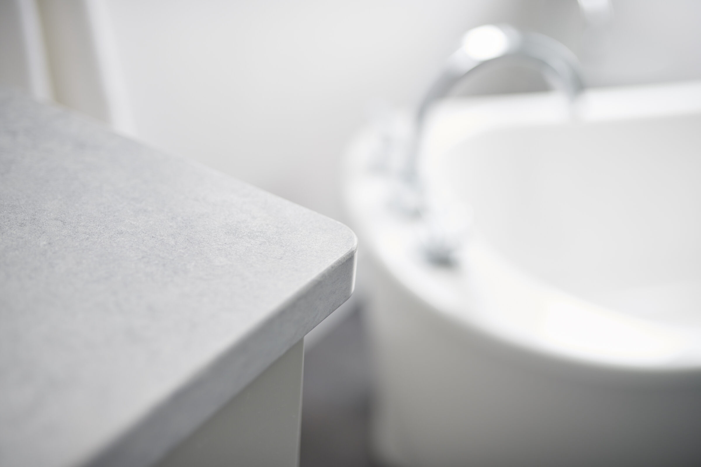 A minimalist bathroom corner with a focused shot on a counter edge, with a blurry background featuring a white sink and a chrome faucet.