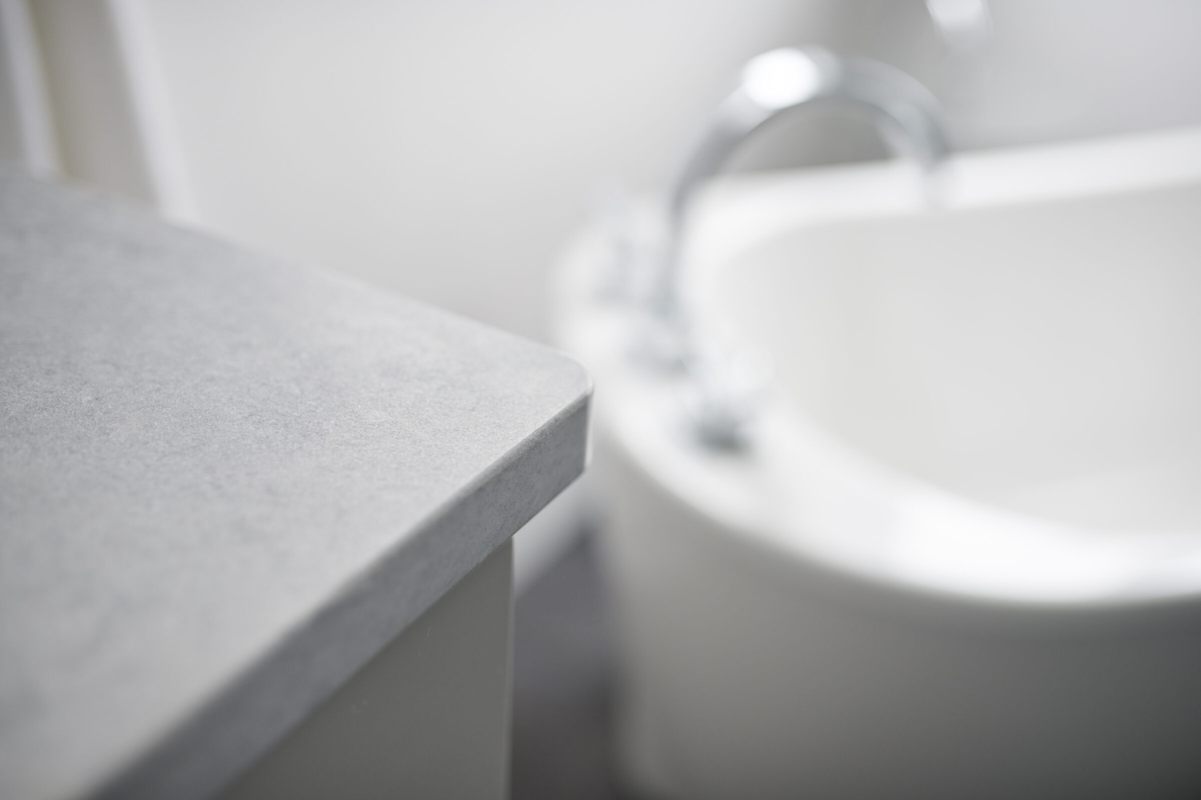A close-up, blurred view of a bathroom interior showing a countertop in focus, and a sink with a faucet in the background out of focus.