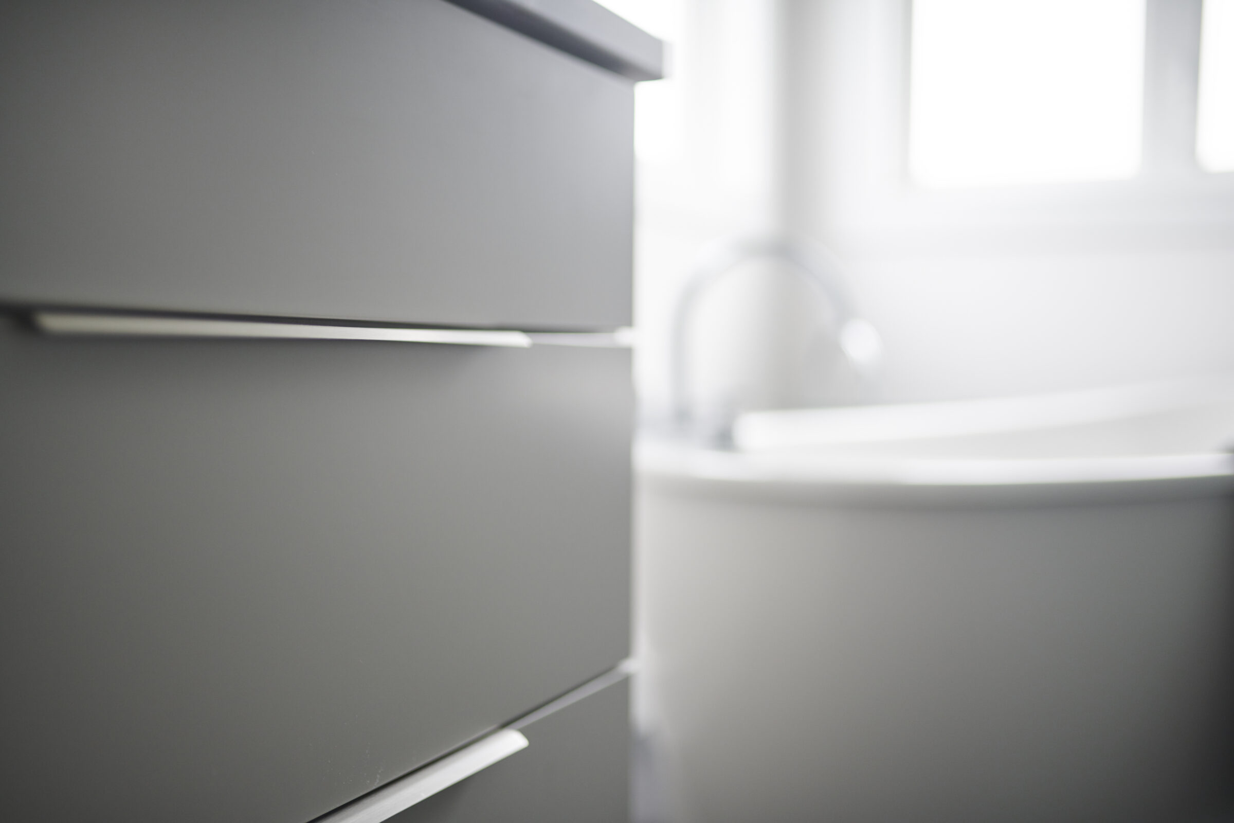 A minimalist bathroom with a focus on a blurred vanity and sink in a bright, airy space with natural light coming from a window.