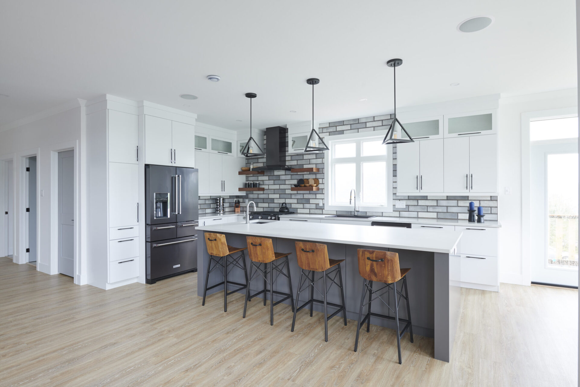 Modern kitchen interior with a central island, wooden stools, black appliances, white cabinetry, gray backsplash tiles, and pendant lighting. No people visible.