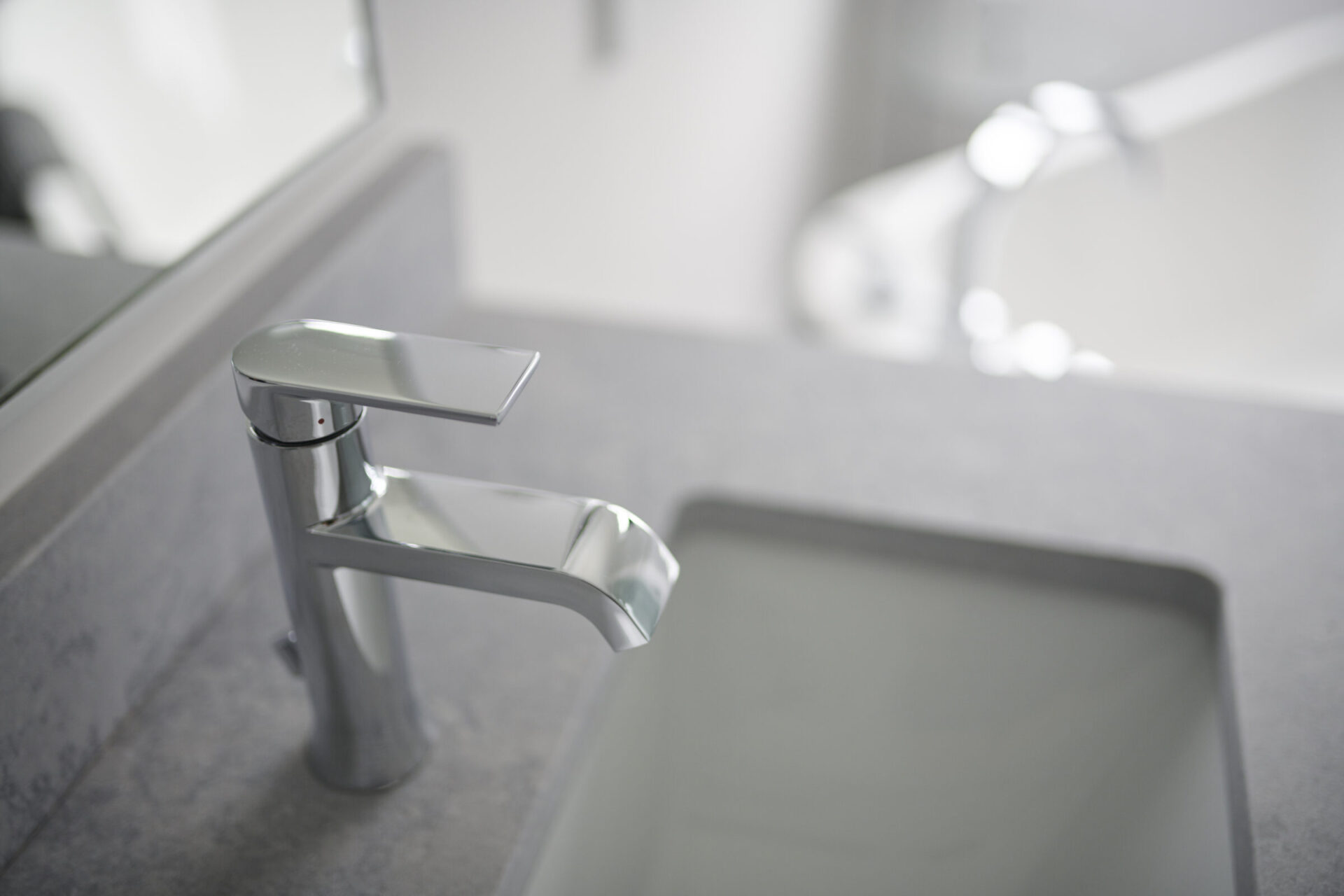 A modern bathroom sink with a sleek, metallic faucet. The countertop is gray, and the blurred reflection in the mirror is seen in the background.