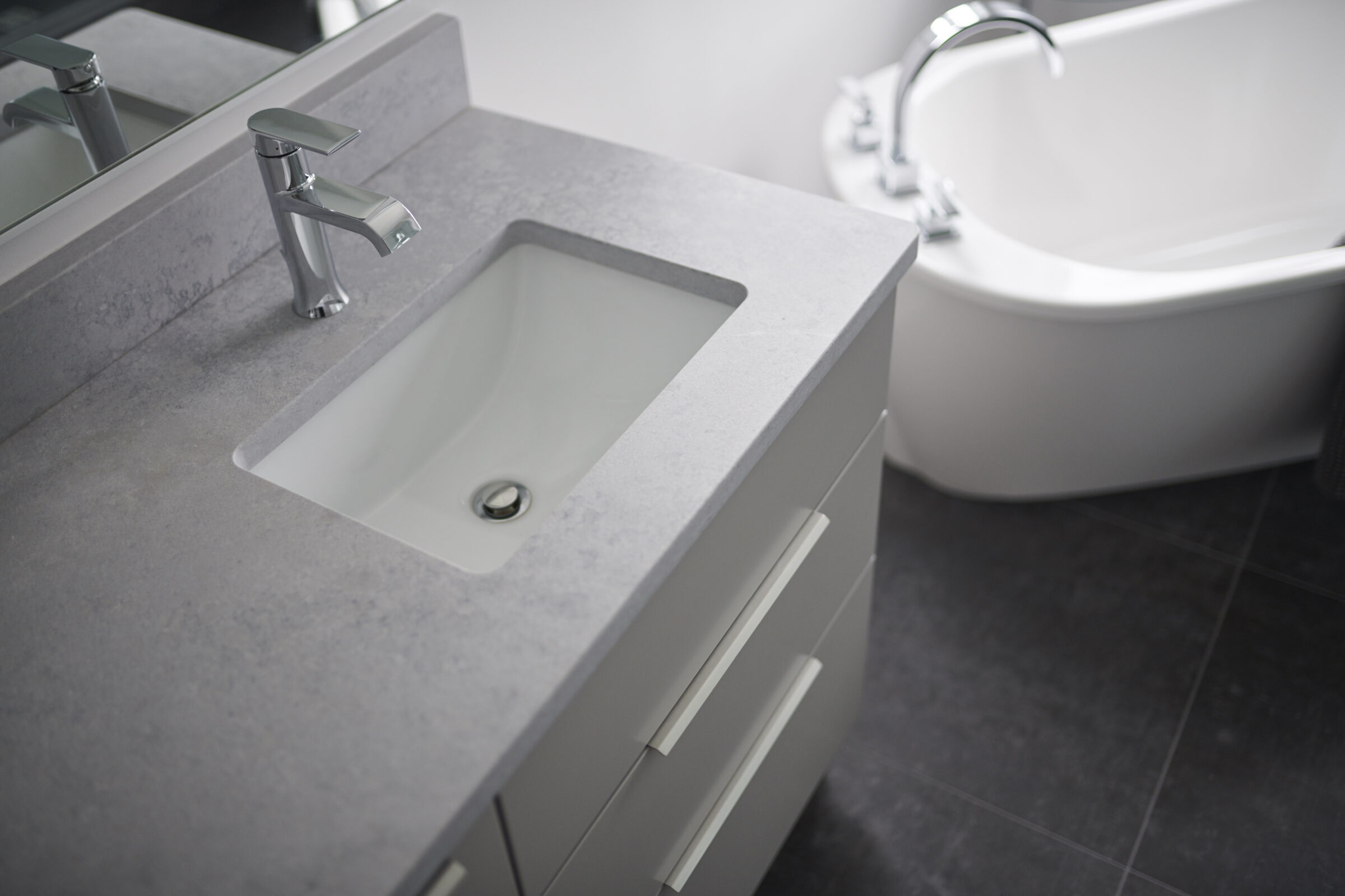 A modern bathroom with a rectangular basin, chrome faucet, and mirror above a grey vanity, with a white bathtub partially visible in the background.
