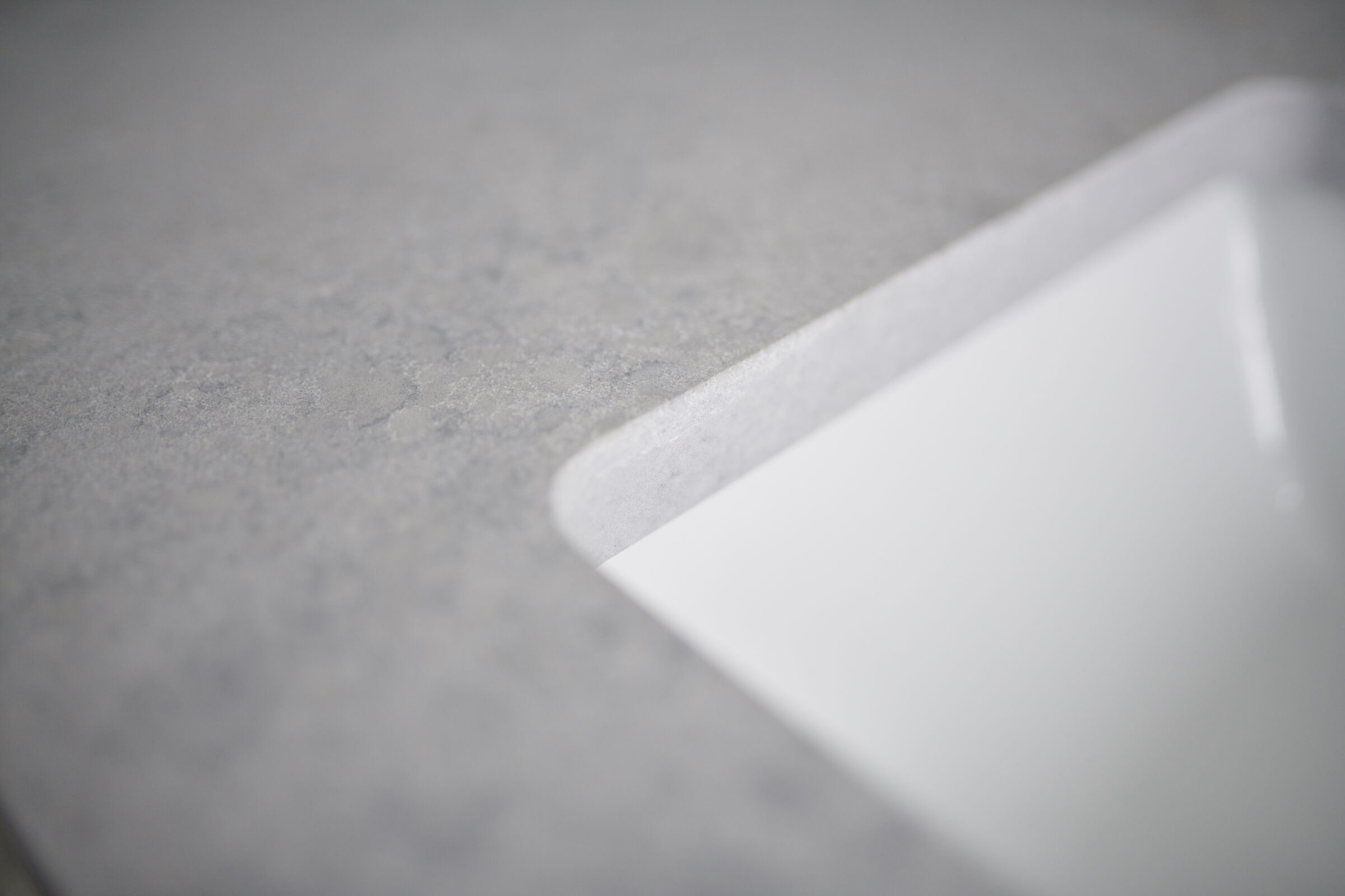 A close-up shot of a smooth, grey surface with the corner of a white object, possibly paper, blurred in the foreground, suggesting simplicity and minimalism.