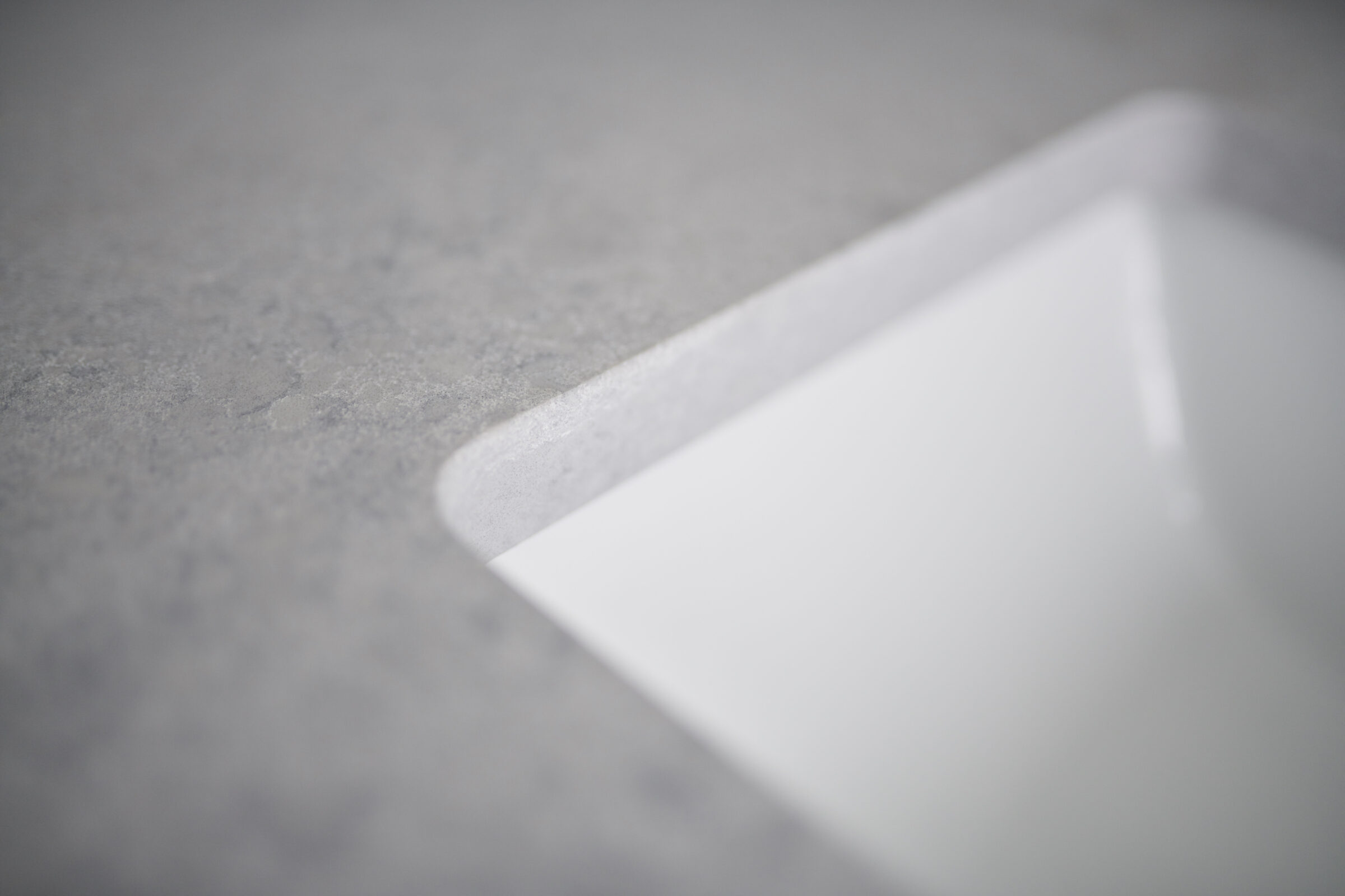 This image shows a close-up of a corner of a white sheet or card on a textured grey surface. The focus is soft, with a shallow depth of field.