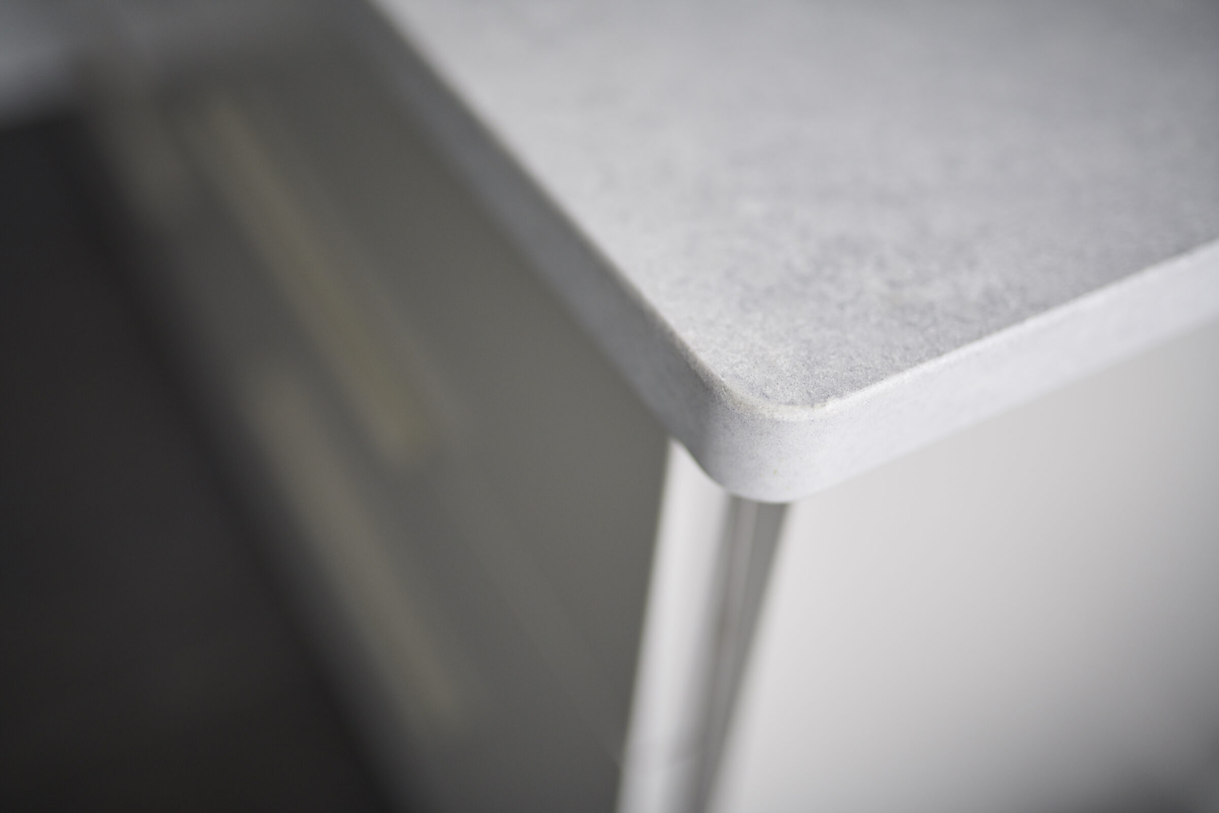 A close-up of a corner of a minimalist gray countertop with soft-focus on the background revealing blurred cabinet structures. The scene feels clean and modern.