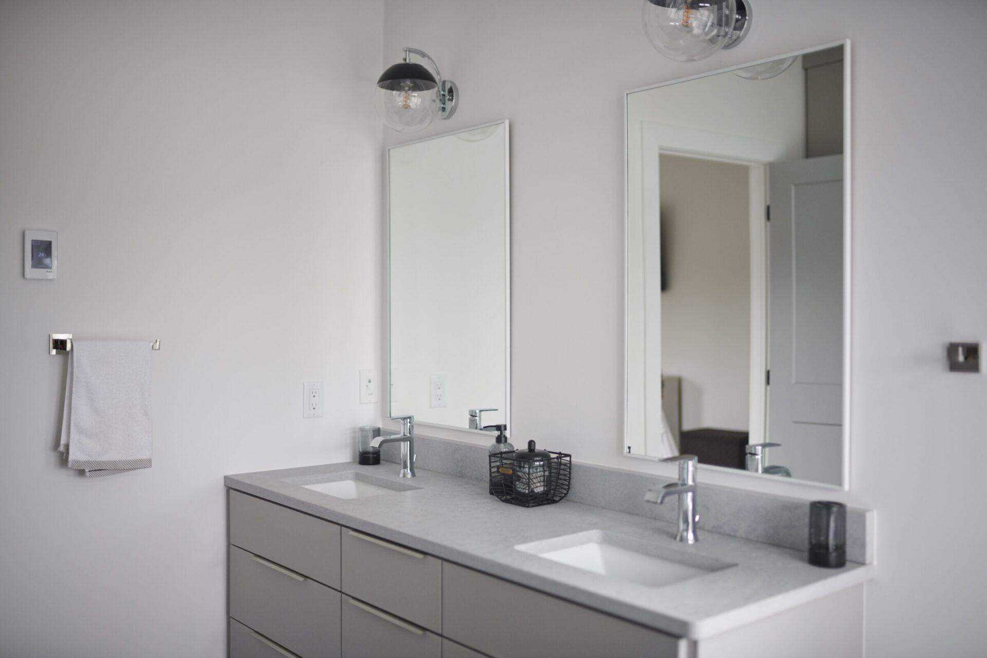 A modern bathroom with a double vanity, two mirrors, globe-style light fixtures, a towel on a rack, and minimalist decor against a neutral palette.