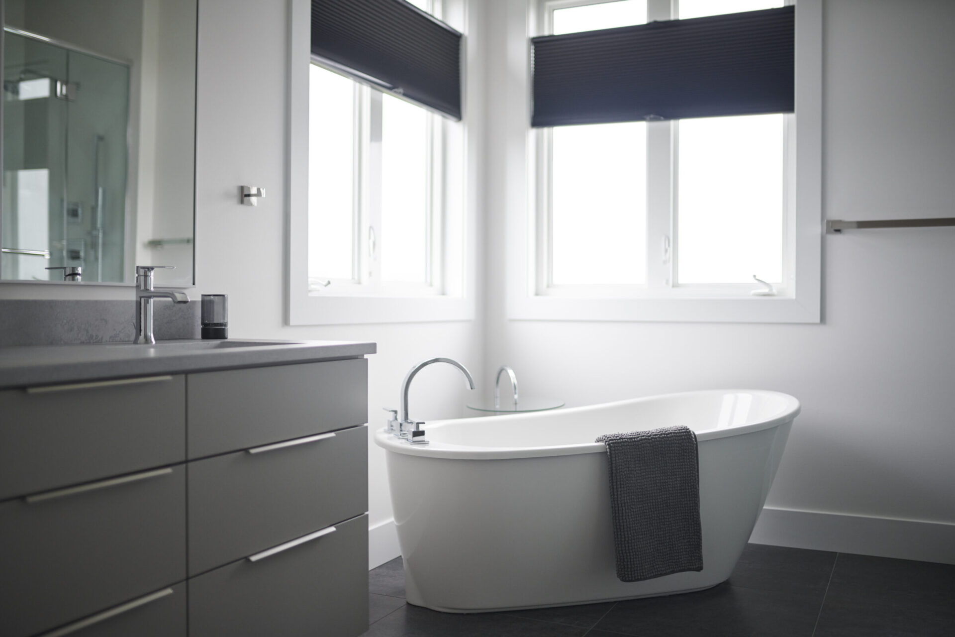 A modern bathroom with a freestanding bathtub, double sink vanity, dark grey floor, and dark blinds on the windows, in a clean, minimalist style.