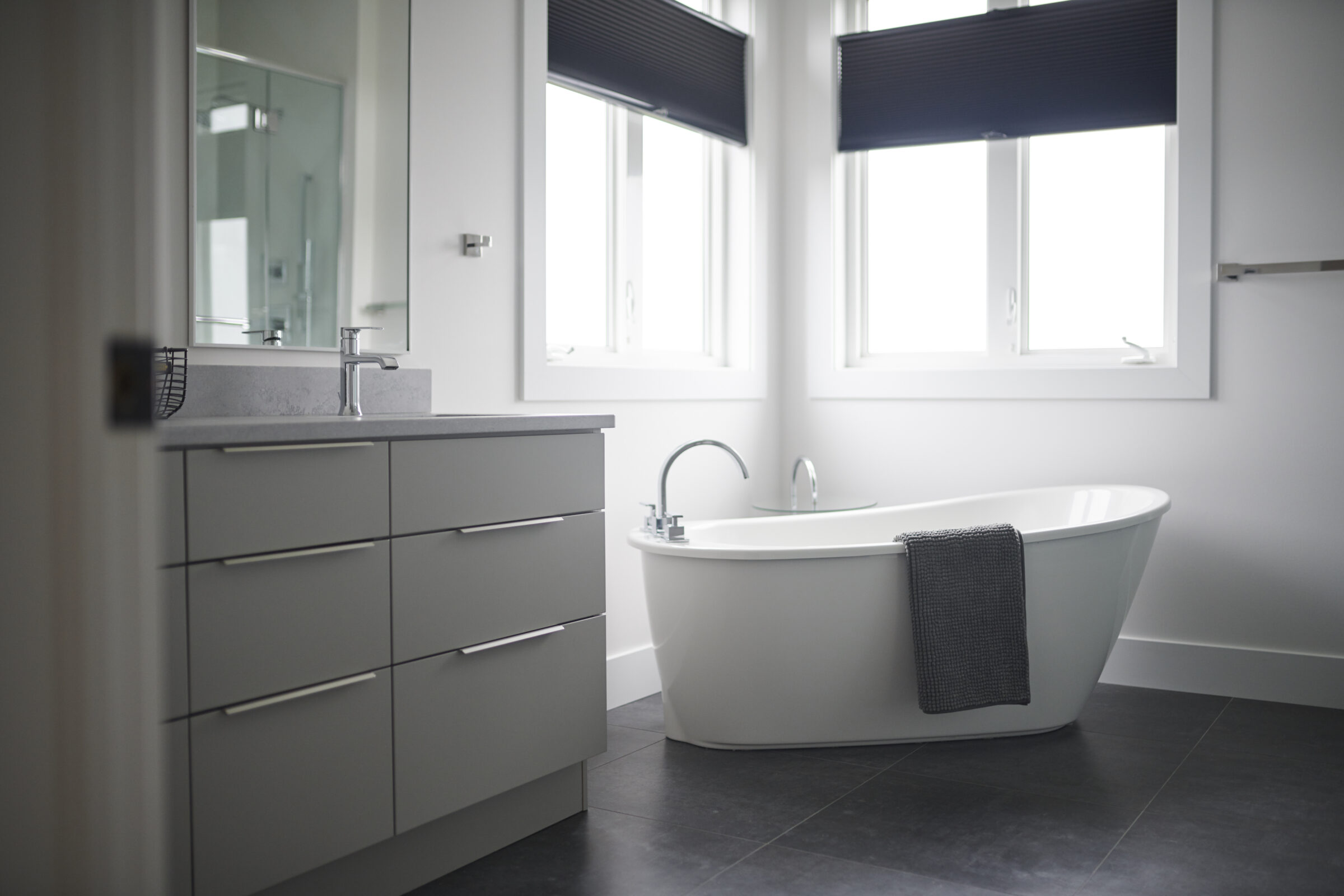 A modern bathroom with a freestanding tub, gray vanity, two windows with dark blinds, and sleek, minimalist design elements. No people are visible.