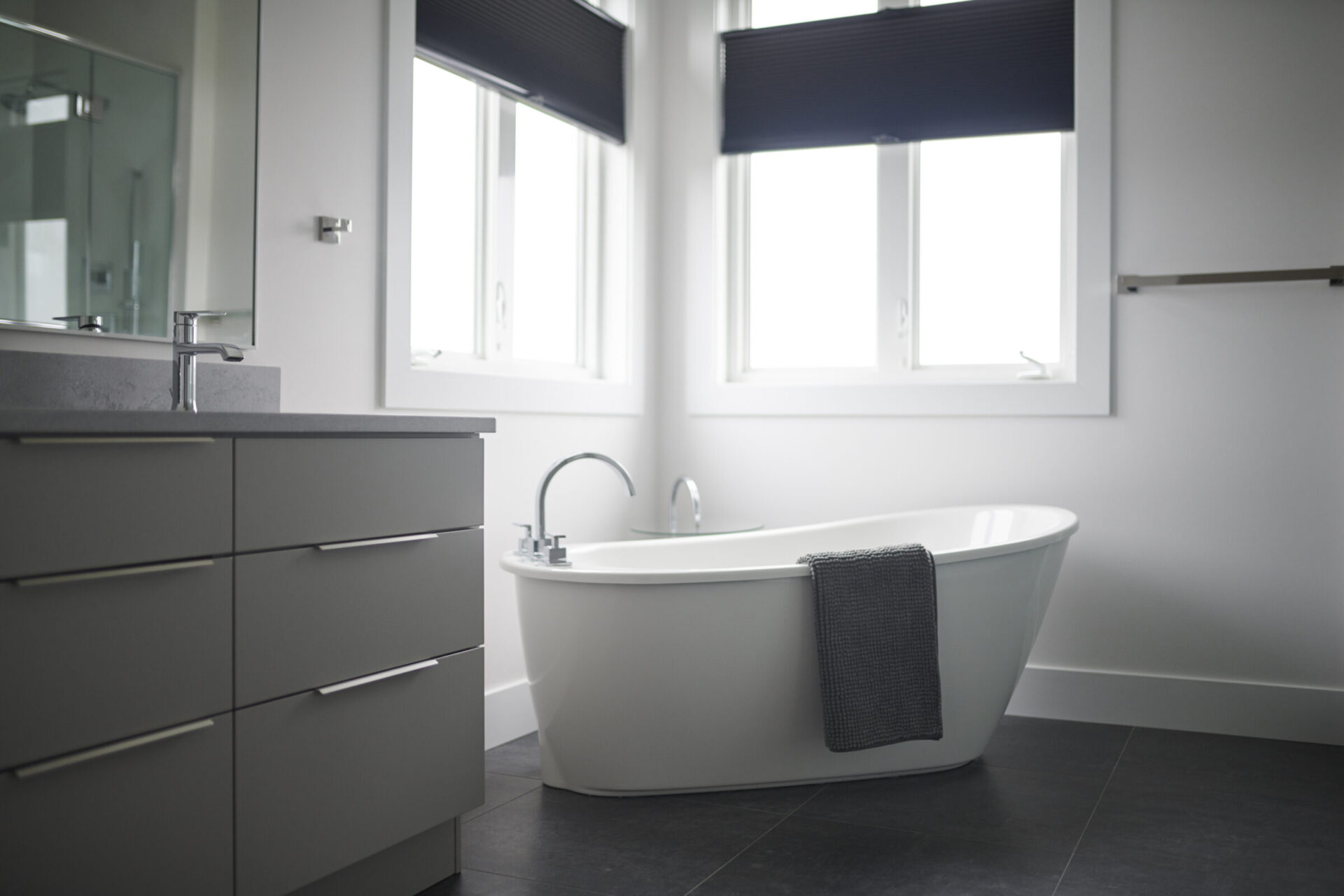 A modern bathroom interior featuring a freestanding bathtub, a vanity with a countertop sink, dark floor tiles, and windows with rolled-up blinds.